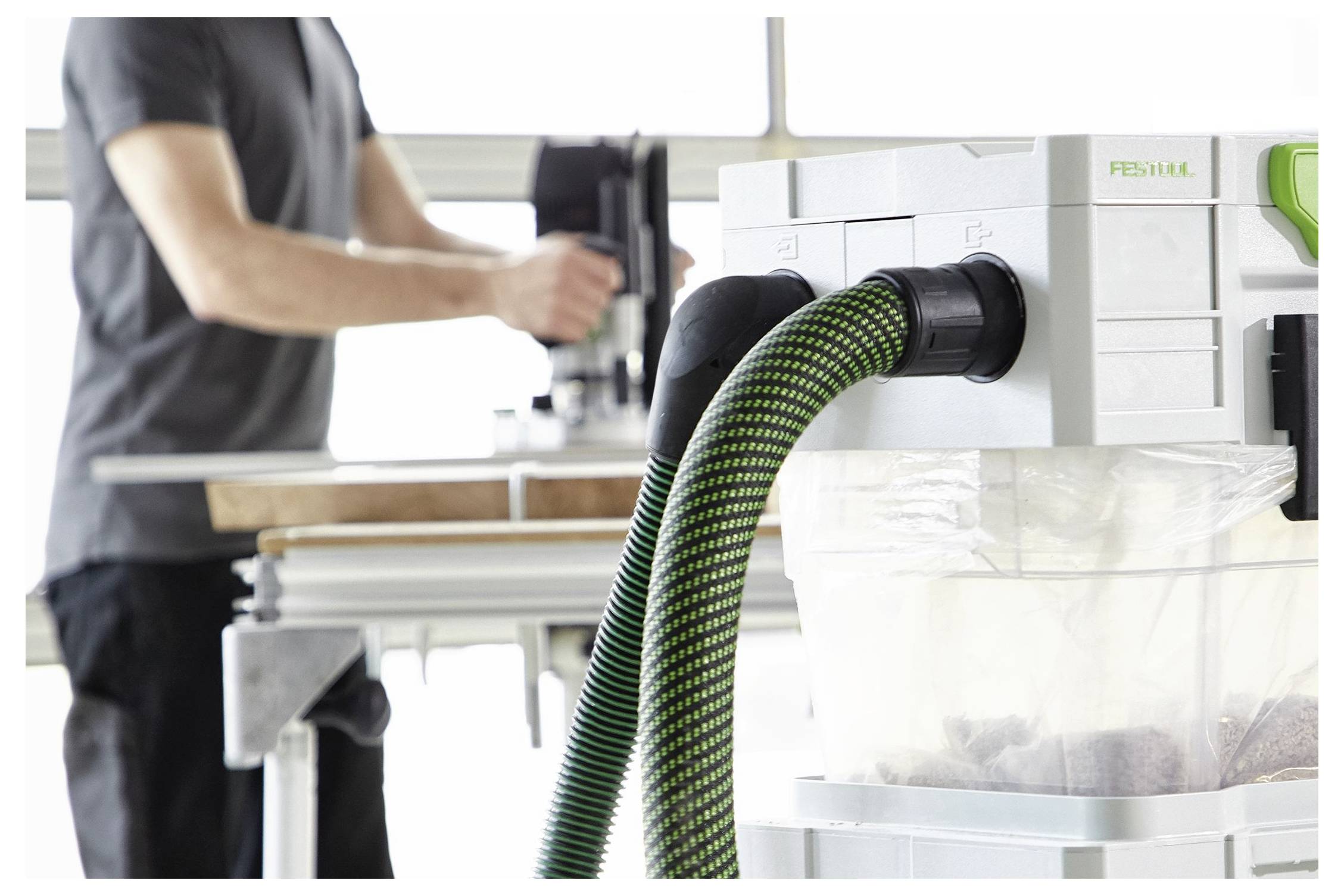 Una persona utilizza attrezzi elettrici in un banco da lavoro; un sistema di aspirazione con un tubo verde cattura i detriti, enfatizzando la pulizia e l'efficienza.