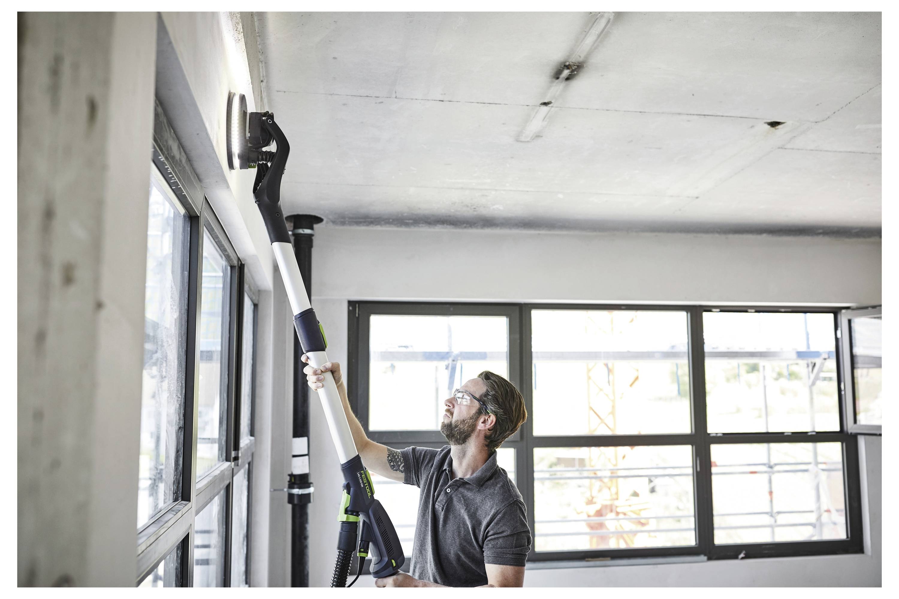 Un uomo utilizza un attrezzo elettrico con manico lungo per lisciare il soffitto in una stanza luminosa con grandi finestre.