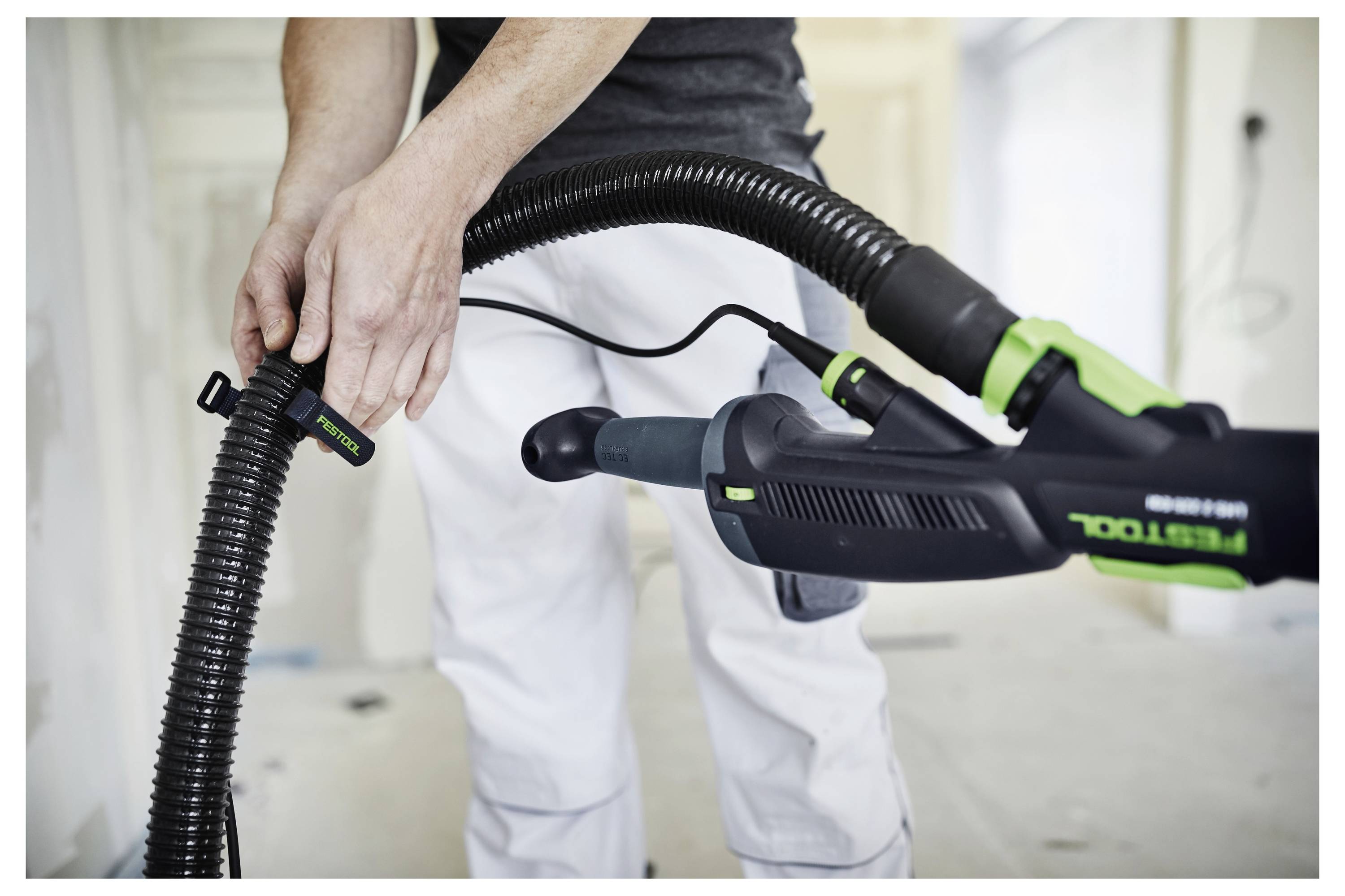 Una persona collega un tubo dell'aspirapolvere a un utensile elettrico verde e nero all'interno di una stanza parzialmente ristrutturata, suggerendo un'attività di pulizia.