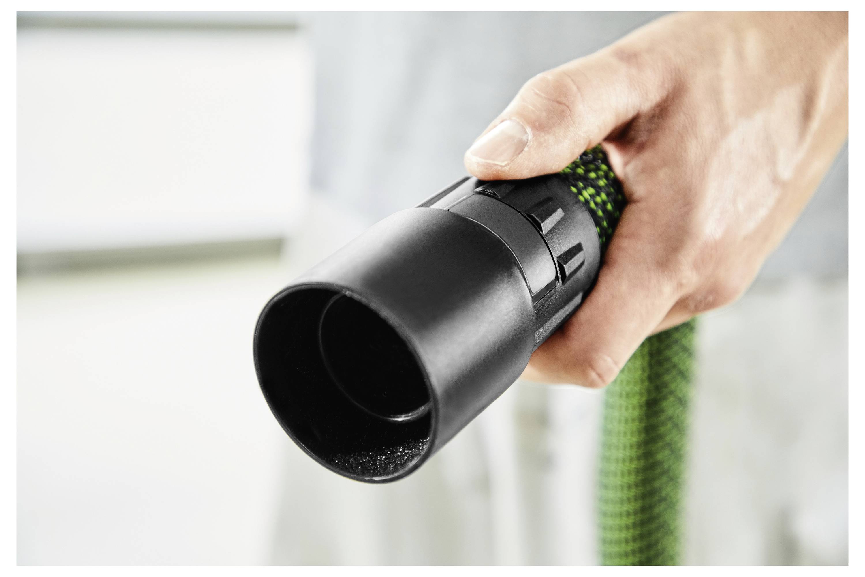 Una mano afferra un tubo da giardino flessibile verde e nero, dirigendo l'ugello spray collegato in avanti. Lo sfondo è sfocato.