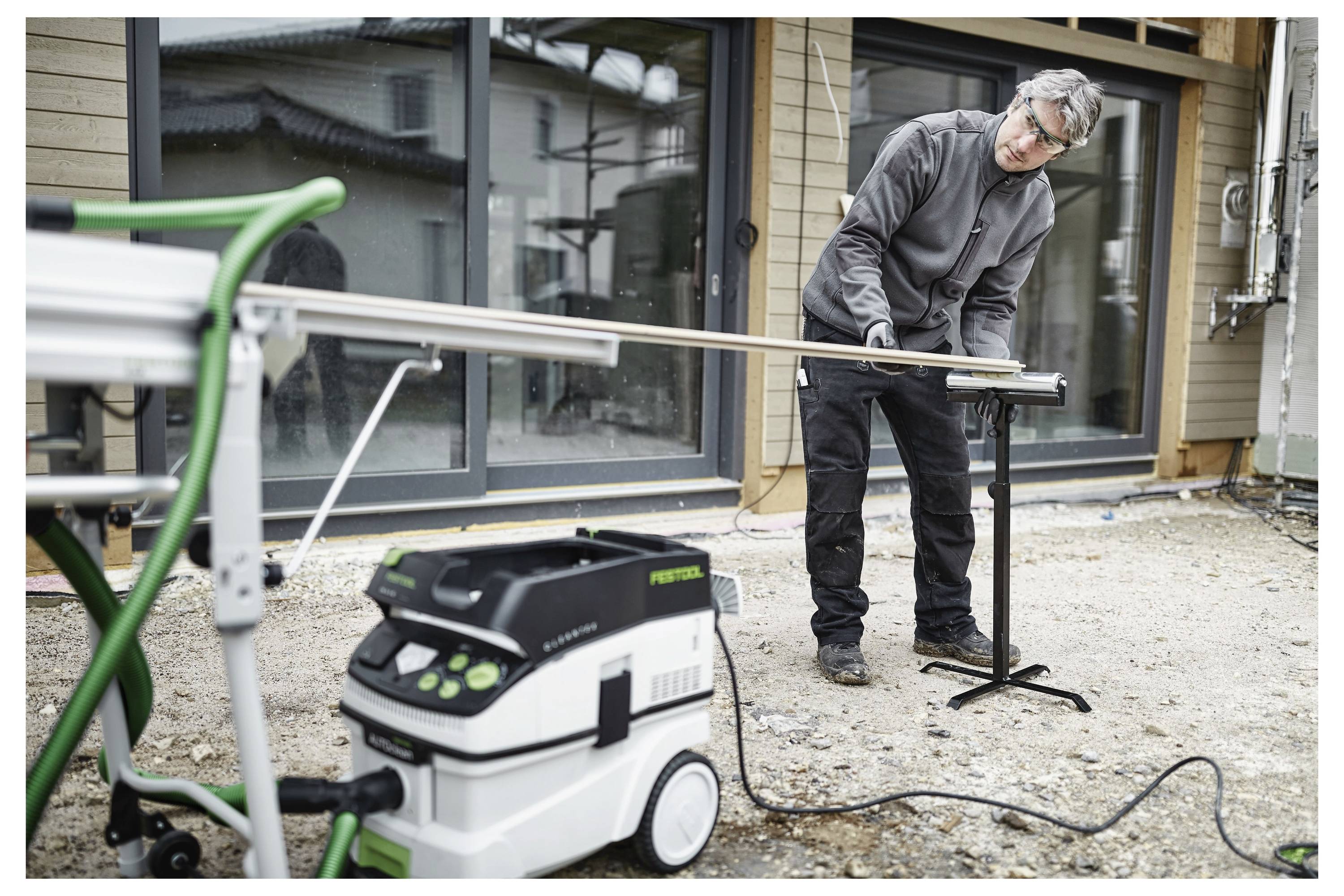 Una persona taglia un lungo pezzo di legno su un cantiere utilizzando una sega e un aspiratore di polvere, stando in piedi davanti a un edificio in ristrutturazione.
