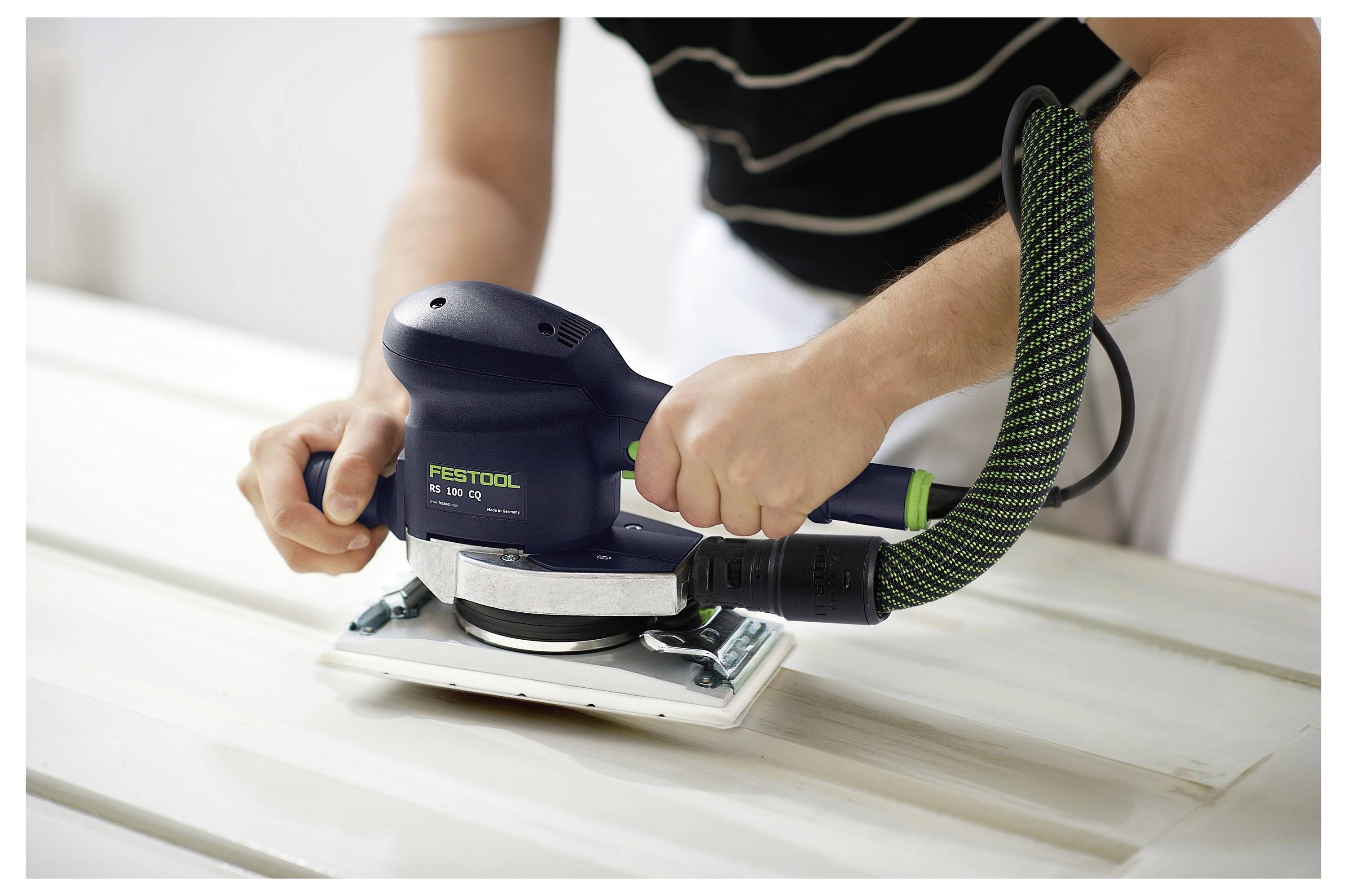 Una persona che utilizza una levigatrice elettrica per lisciare una superficie di legno, tenendo il dispositivo con entrambe le mani. La levigatrice è collegata a un tubo.