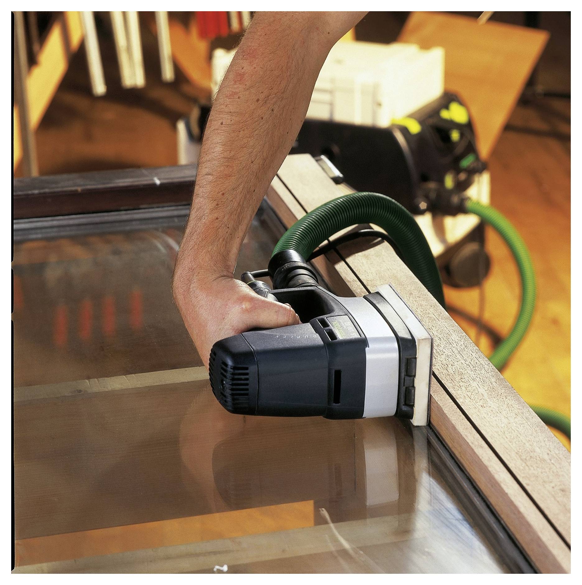 Una persona utilizza una levigatrice elettrica su una cornice di legno, con un tubo verde collegato, in un'ambientazione di officina.