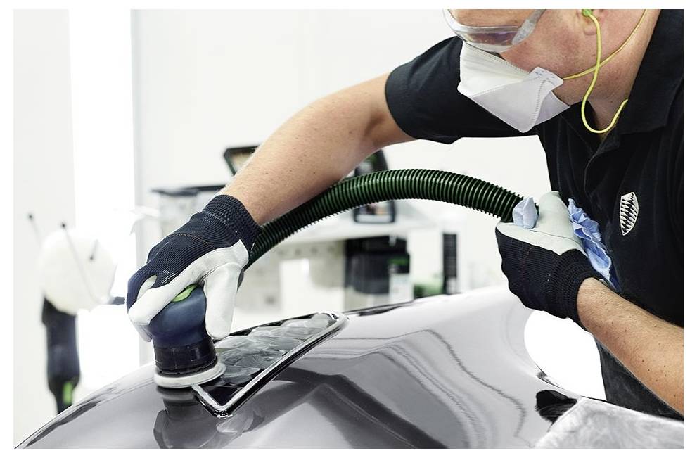 'Una persona che indossa equipaggiamento protettivo utilizza una lucidatrice sul cofano di un'auto, evidenziando la lucidatura della superficie e la cura in un'officina automobilistica.'