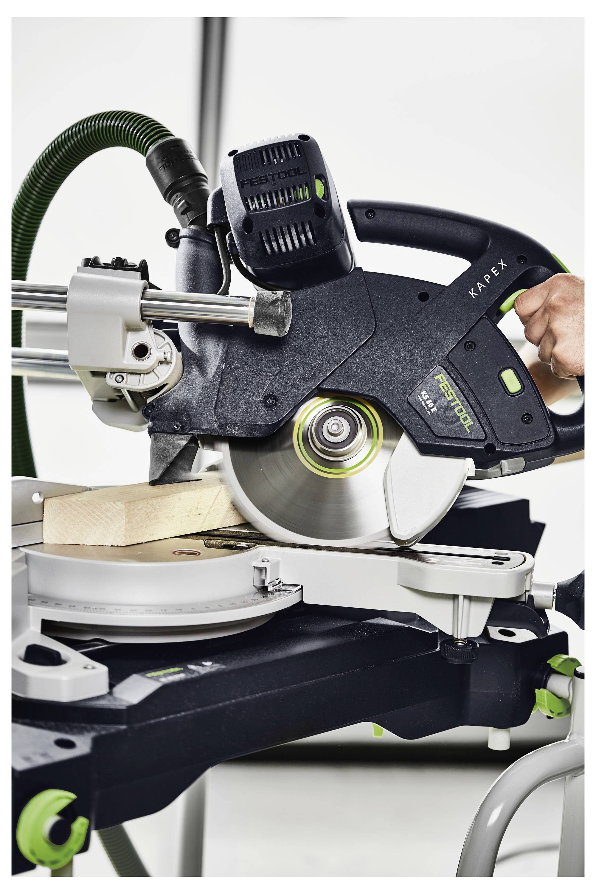Una sega circolare che taglia un pezzo di legno su un banco da lavoro, con un manico visibile per l'uso e un tubo di raccolta della polvere collegato.