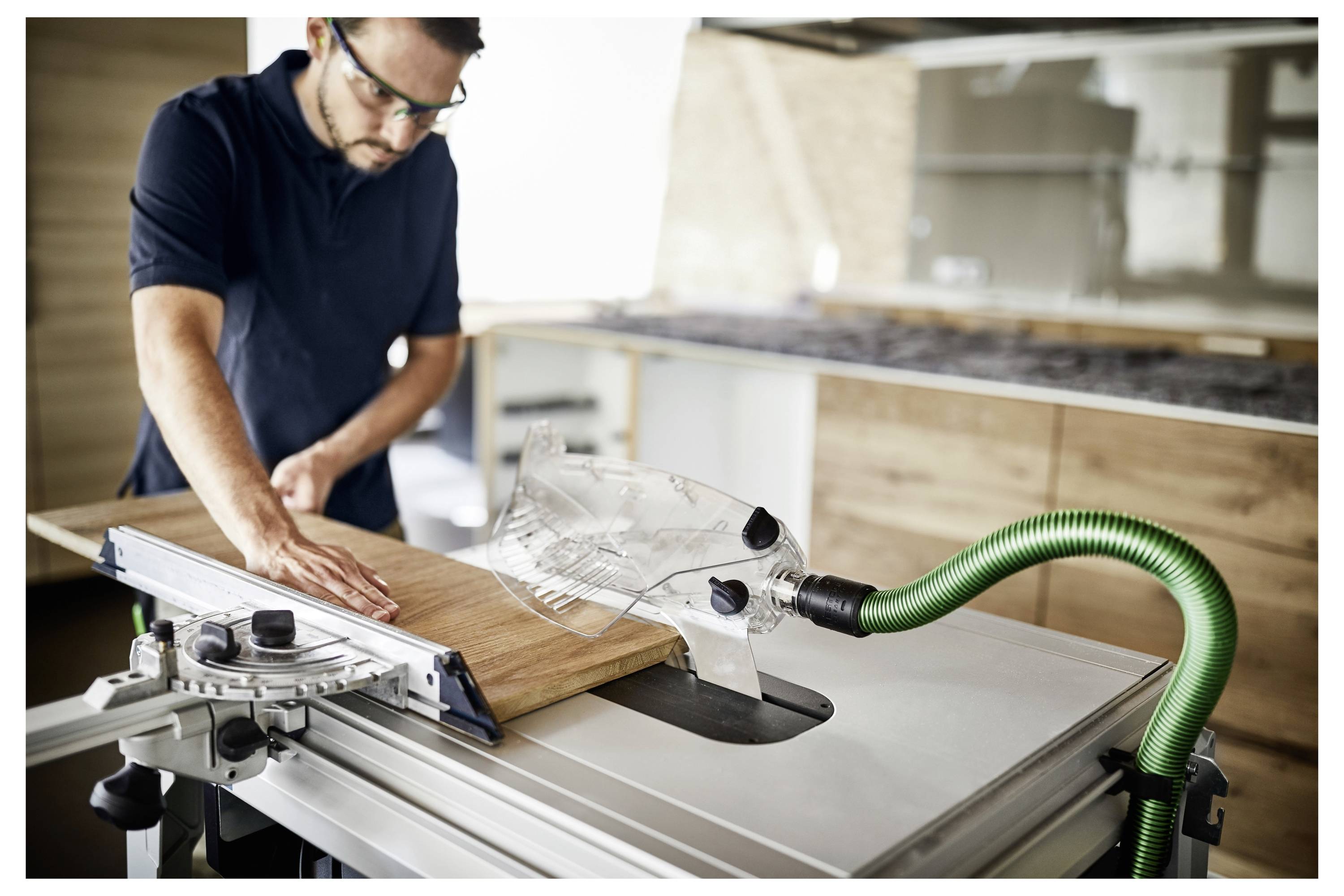 Una persona che utilizza una sega circolare per tagliare un pezzo di legno con un tubo di estrazione della polvere verde collegato, indossando occhiali di sicurezza per la protezione.