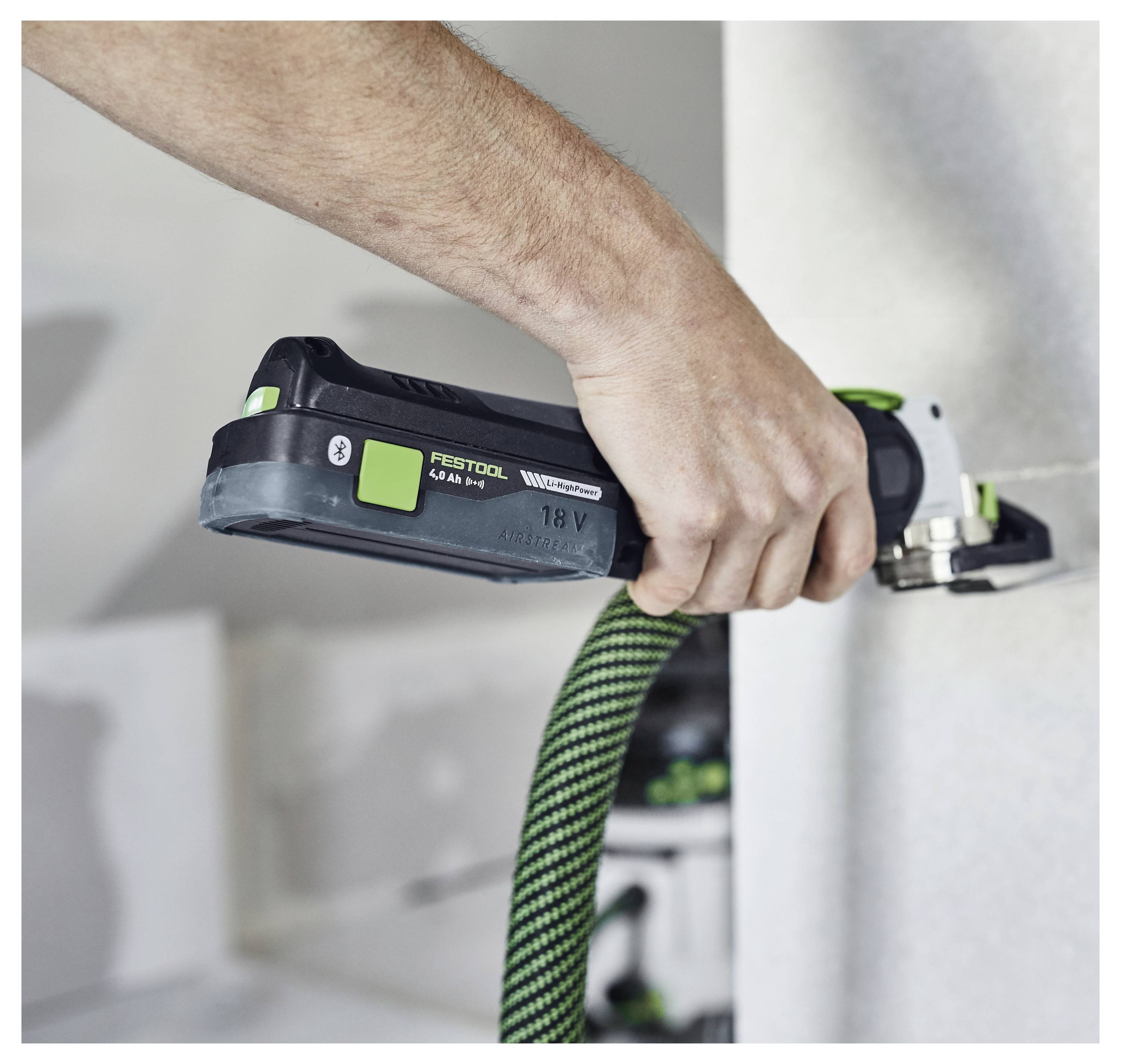 Una persona che utilizza un utensile elettrico di levigatura con manico verde e nero per levigare una superficie di cartongesso. L'utensile è collegato a un tubo.