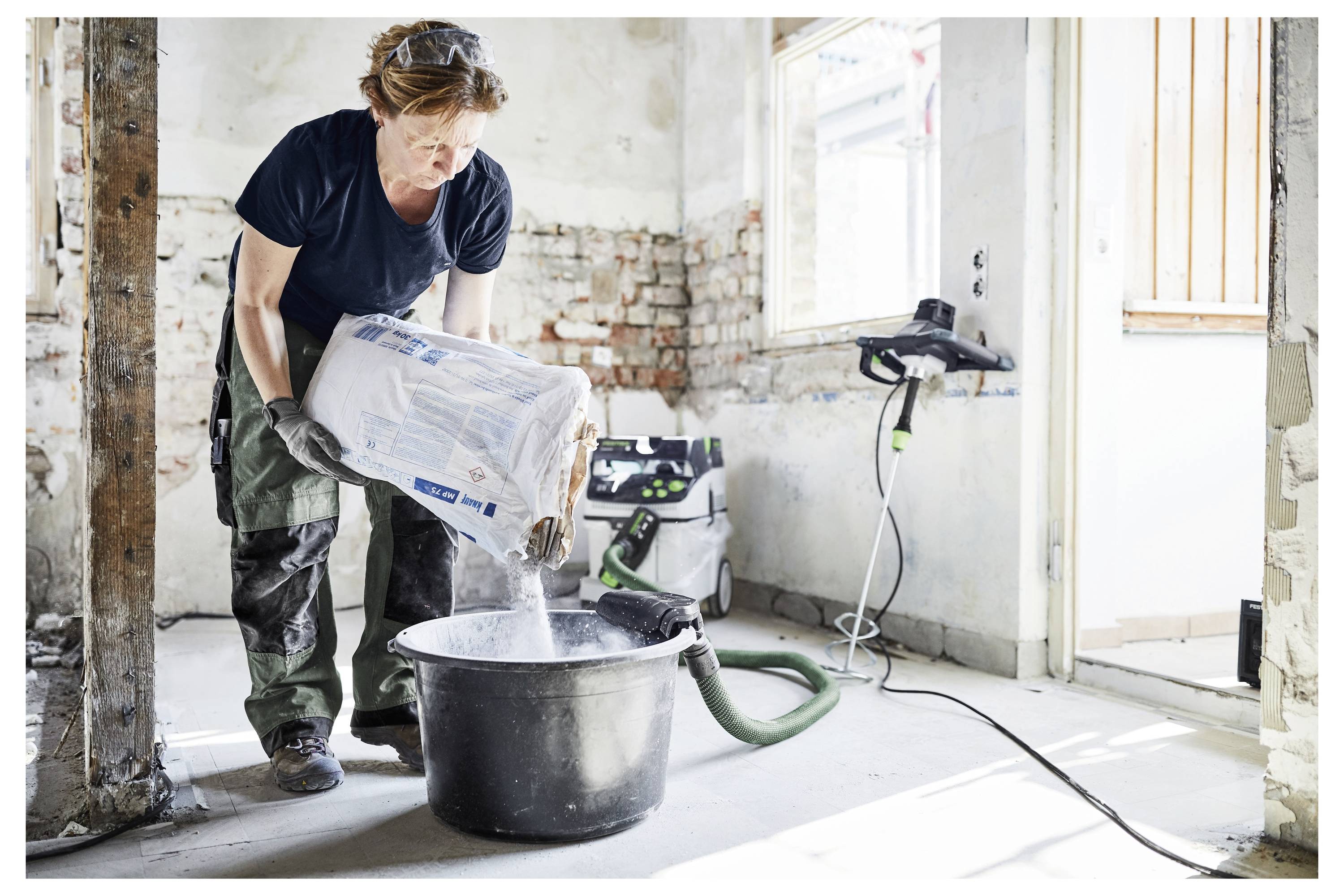 Una persona versa un sacchetto di materiale da costruzione in un secchio nero all'interno di una stanza parzialmente ristrutturata con mattoni a vista e una finestra.