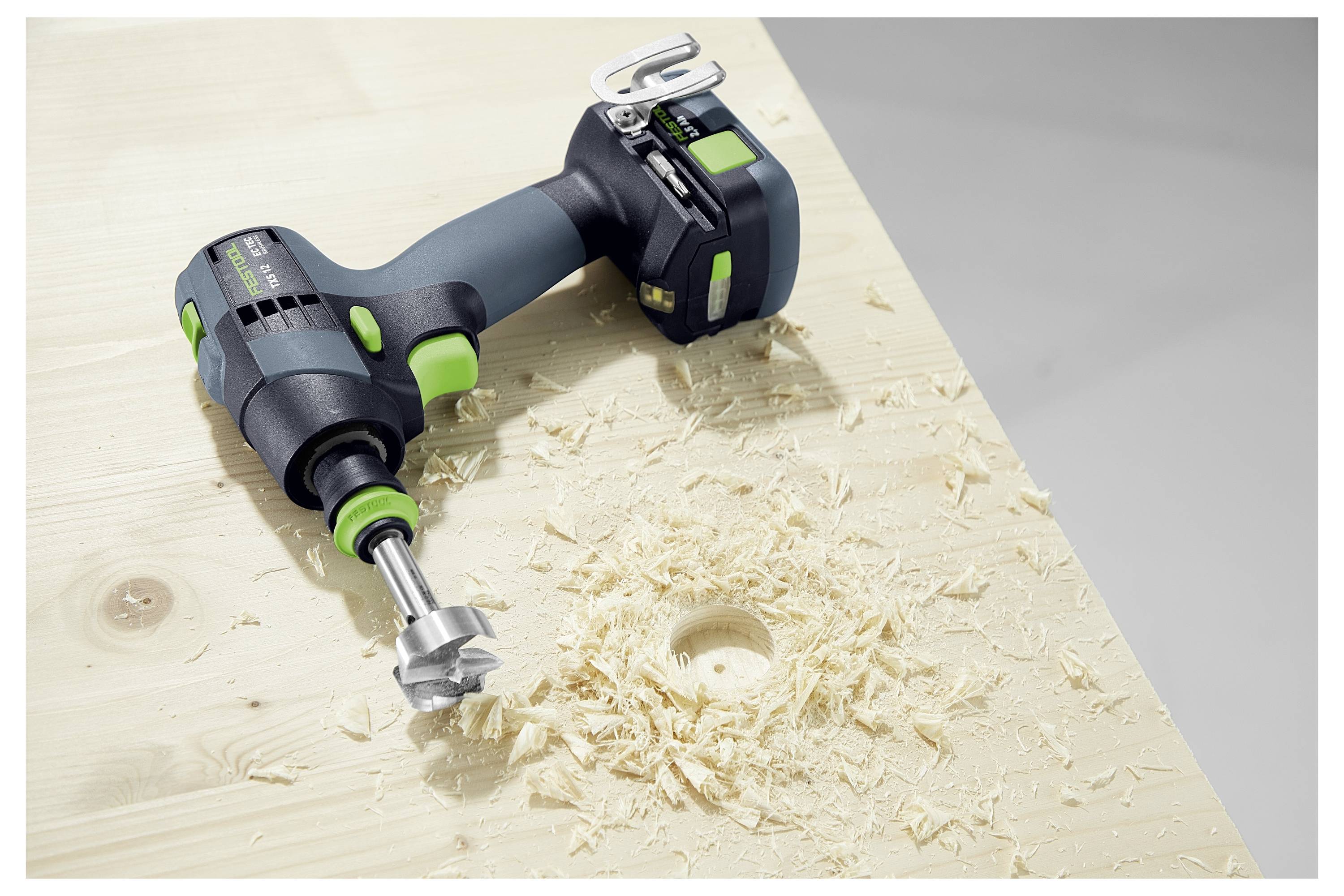 Una trapano a batteria con una punta da trapano attaccata si trova su una superficie di legno leggero con trucioli di legno intorno a un foro trapanato.