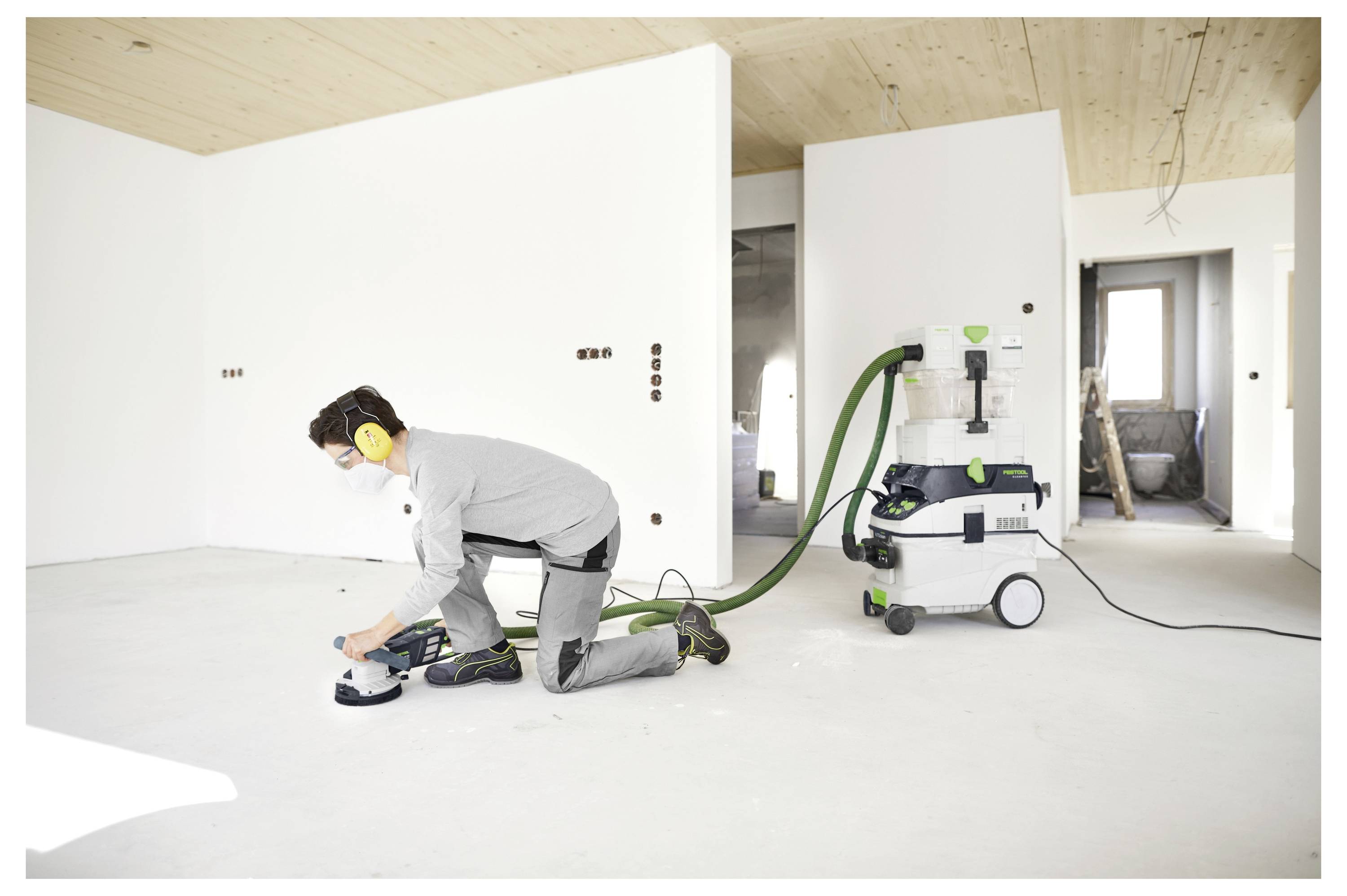 Una persona è in ginocchio sul pavimento mentre utilizza una levigatrice elettrica in una stanza bianca in fase di costruzione. Indossa un equipaggiamento di sicurezza e un estrattore di polvere è collegato.