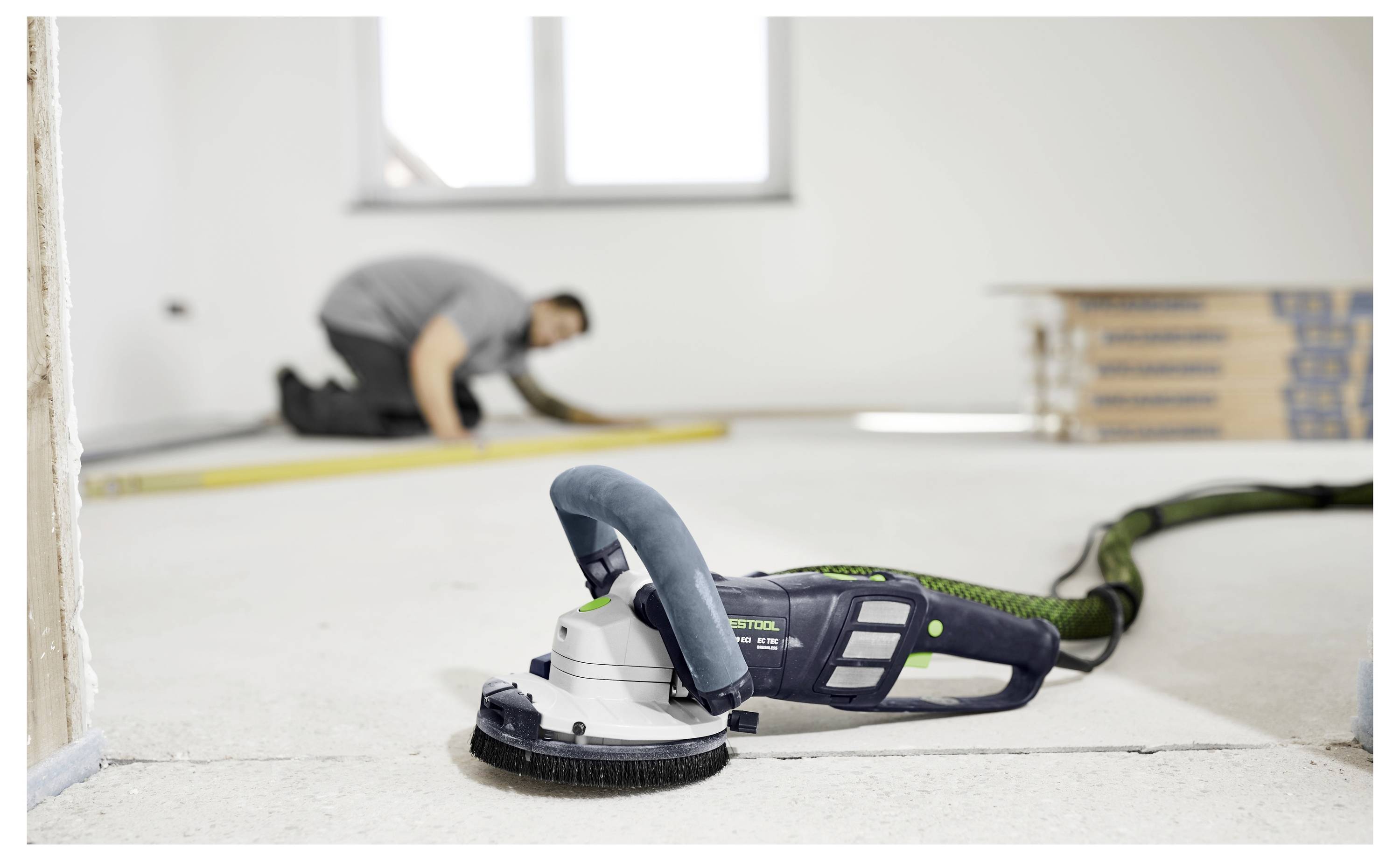 Un operaio edile misura il pavimento sullo sfondo mentre una levigatrice pavimenti professionale giace in primo piano, evidenziando i lavori di ristrutturazione.