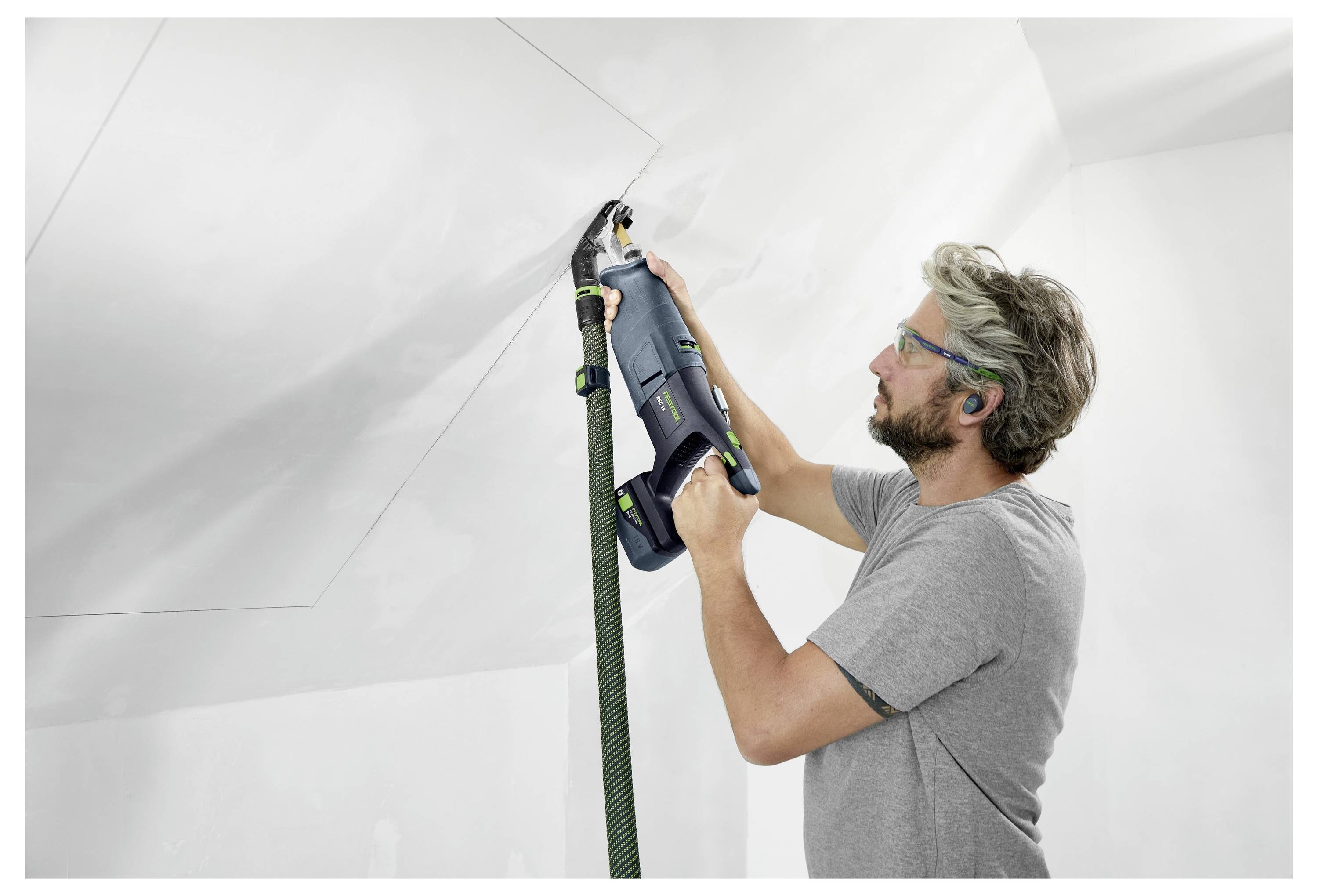 Un uomo che indossa occhiali di sicurezza utilizza un utensile elettrico per carteggiare un soffitto, assicurando una superficie liscia. Tiene il tubo dell'aspirapolvere collegato all'utensile.