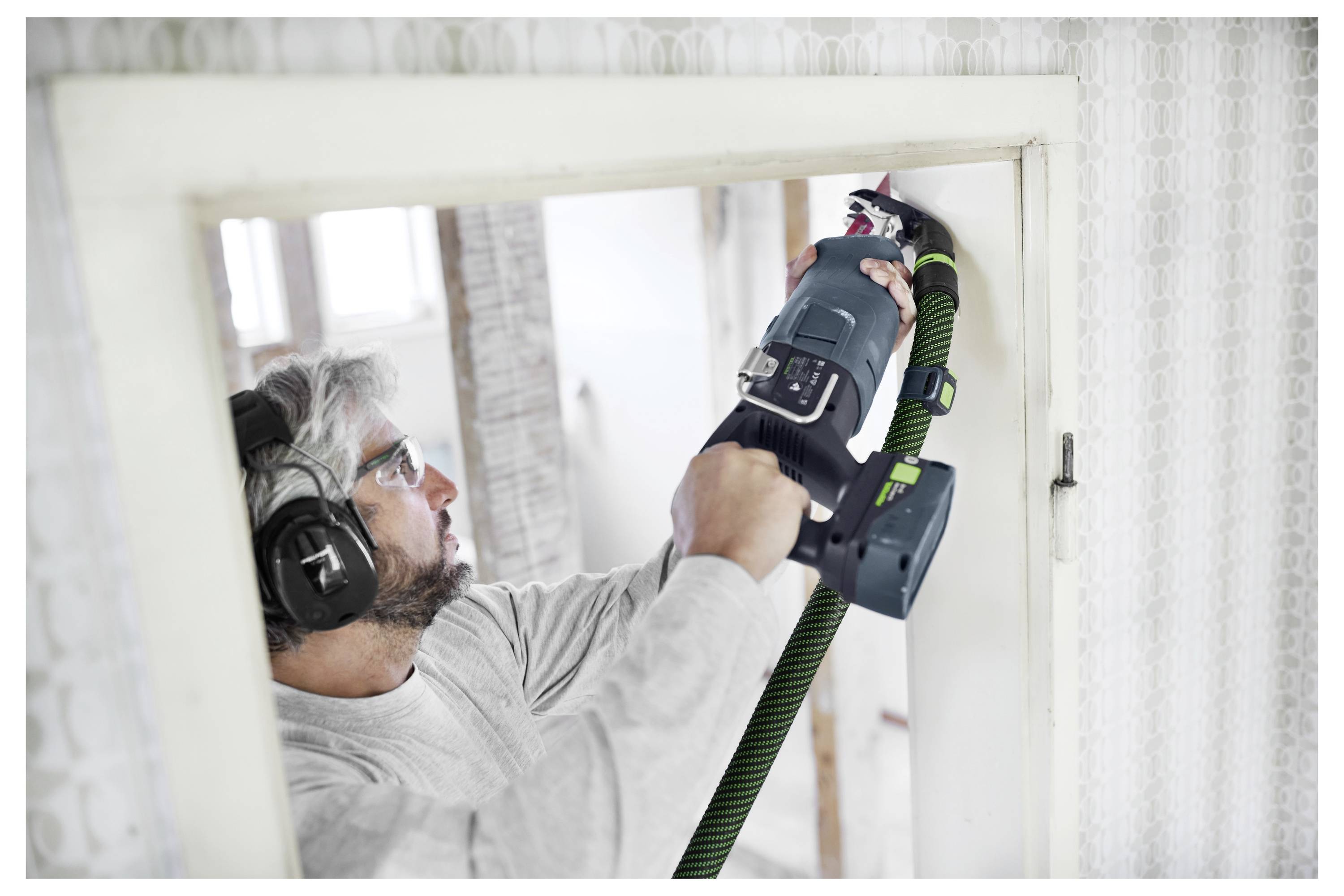 Una persona che indossa occhiali di sicurezza e cuffie antirumore sta utilizzando un utensile elettrico per tagliare attraverso una parete in una stanza luminosa sottoposta a ristrutturazione.