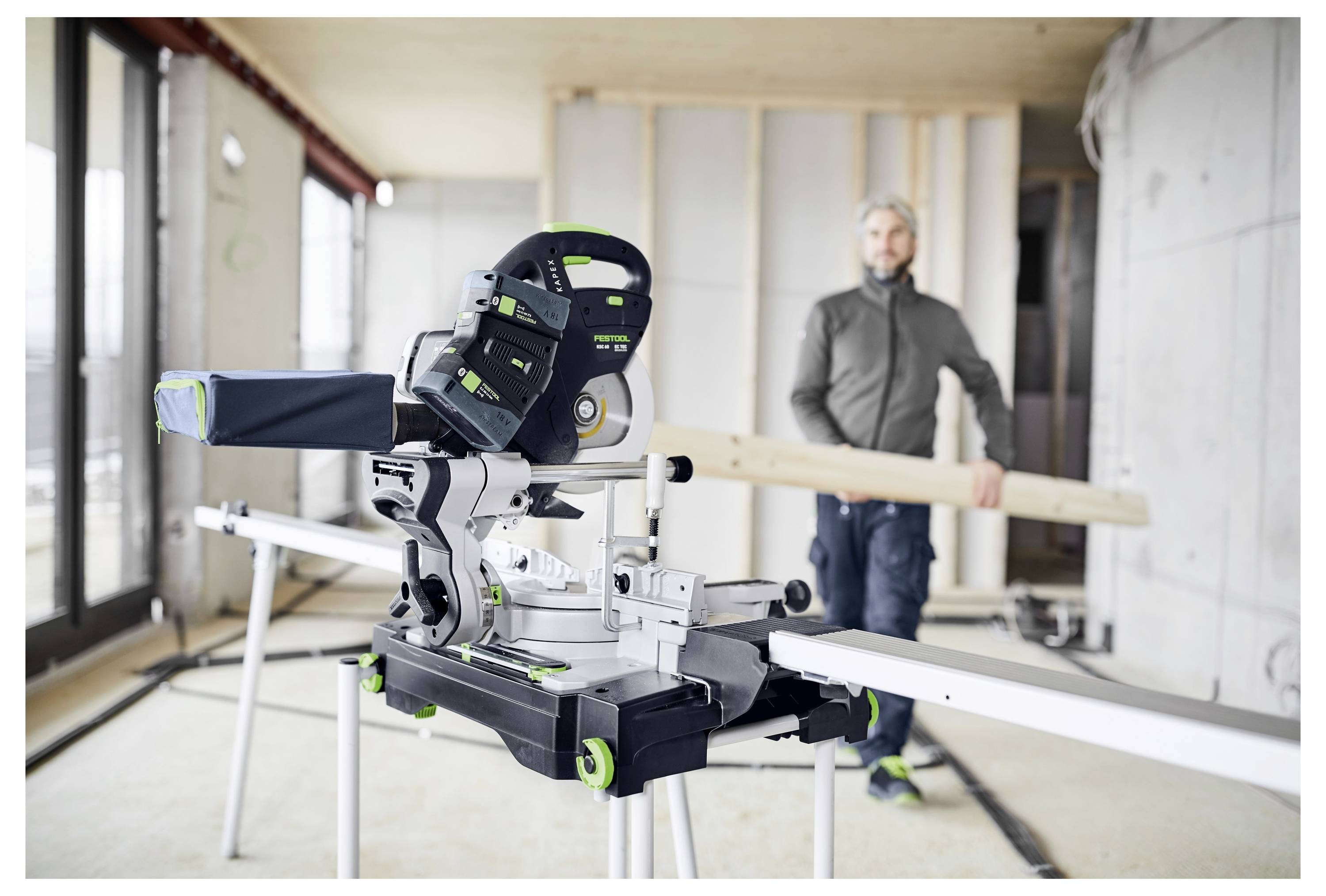 Una persona in una stanza parzialmente costruita che trasporta una trave di legno. In primo piano, una sega a spigolo è posizionata su un banco di lavoro.