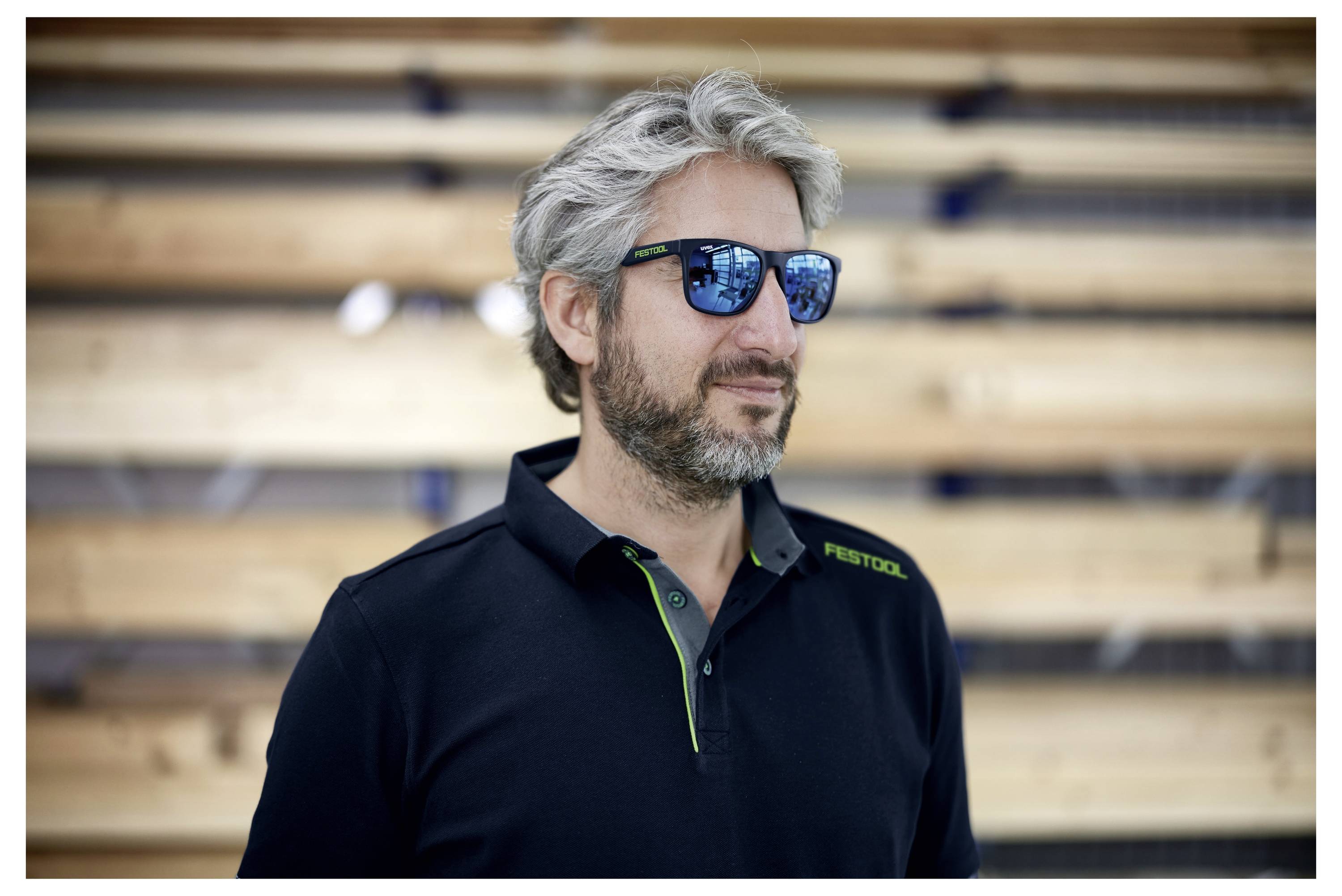 Uomo con capelli grigi e barba indossa occhiali da sole scuri e una maglietta nera in piedi di fronte a uno sfondo con assi di legno orizzontali.