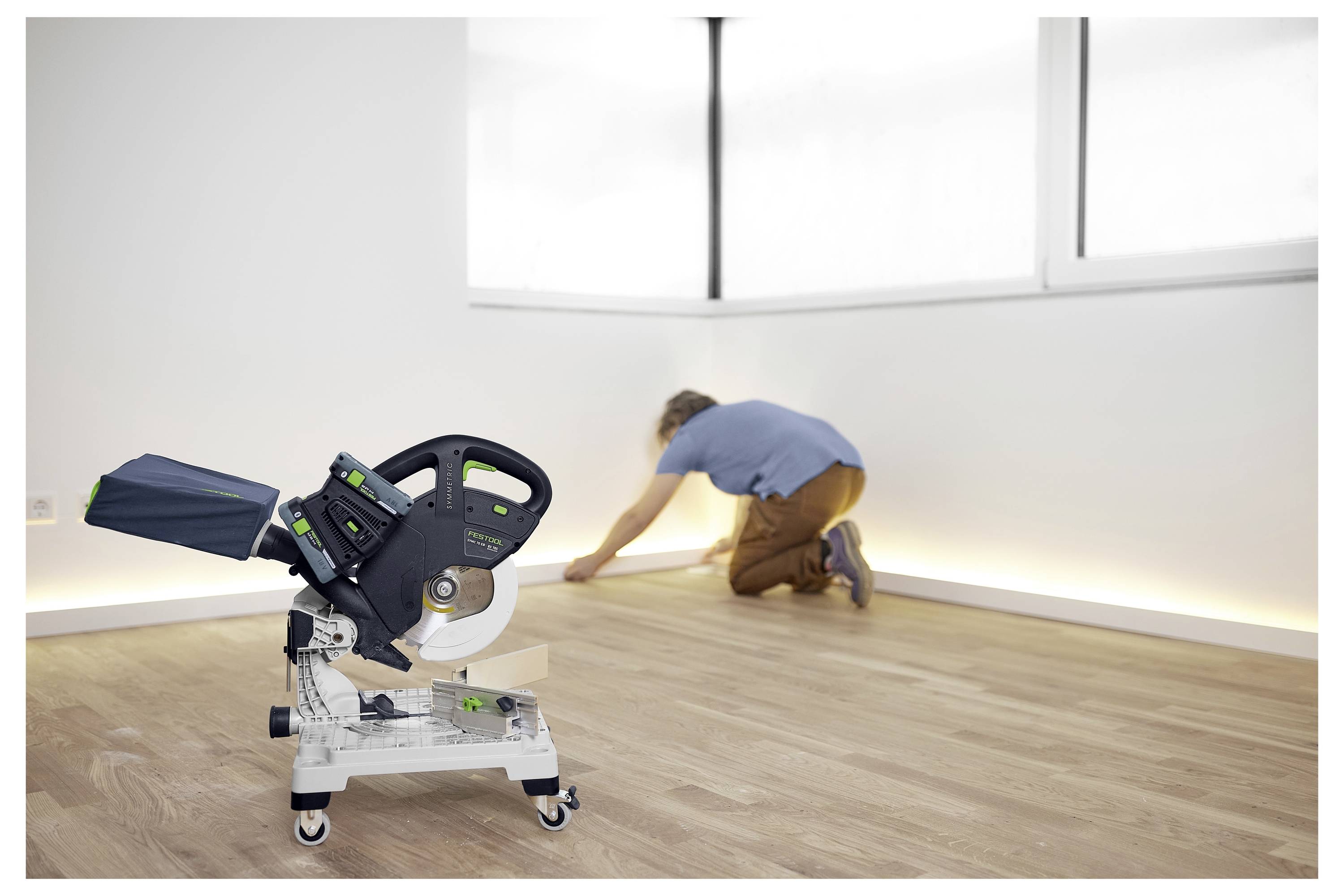 Una sega elettrica è in primo piano su un pavimento di legno. Sullo sfondo, una persona è inginocchiata mentre installa battiscopa in una stanza ben illuminata.
