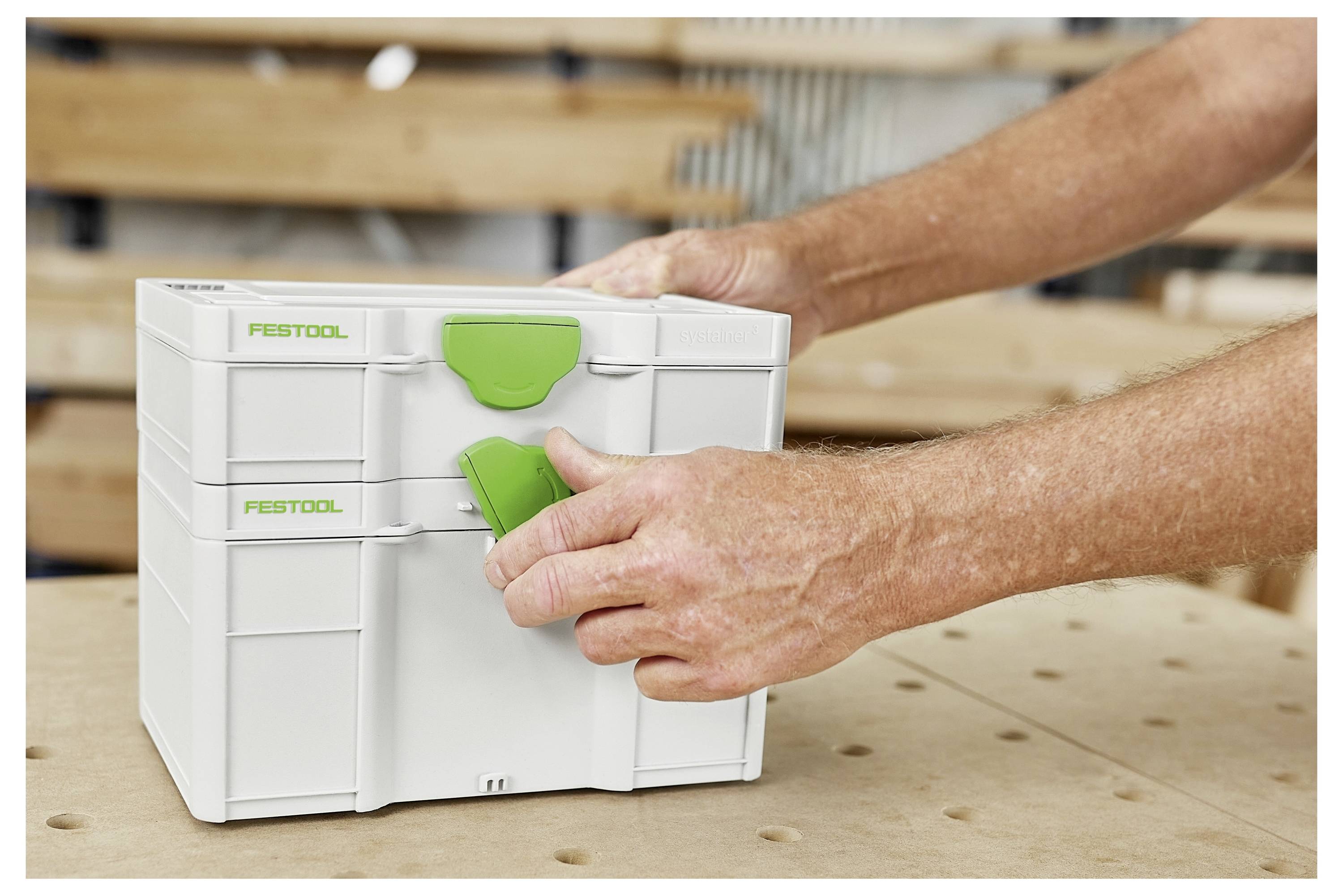 Due mani stanno aprendo una scatola di stoccaggio bianca Festool Systainer con fermi verdi su un banco da lavoro.