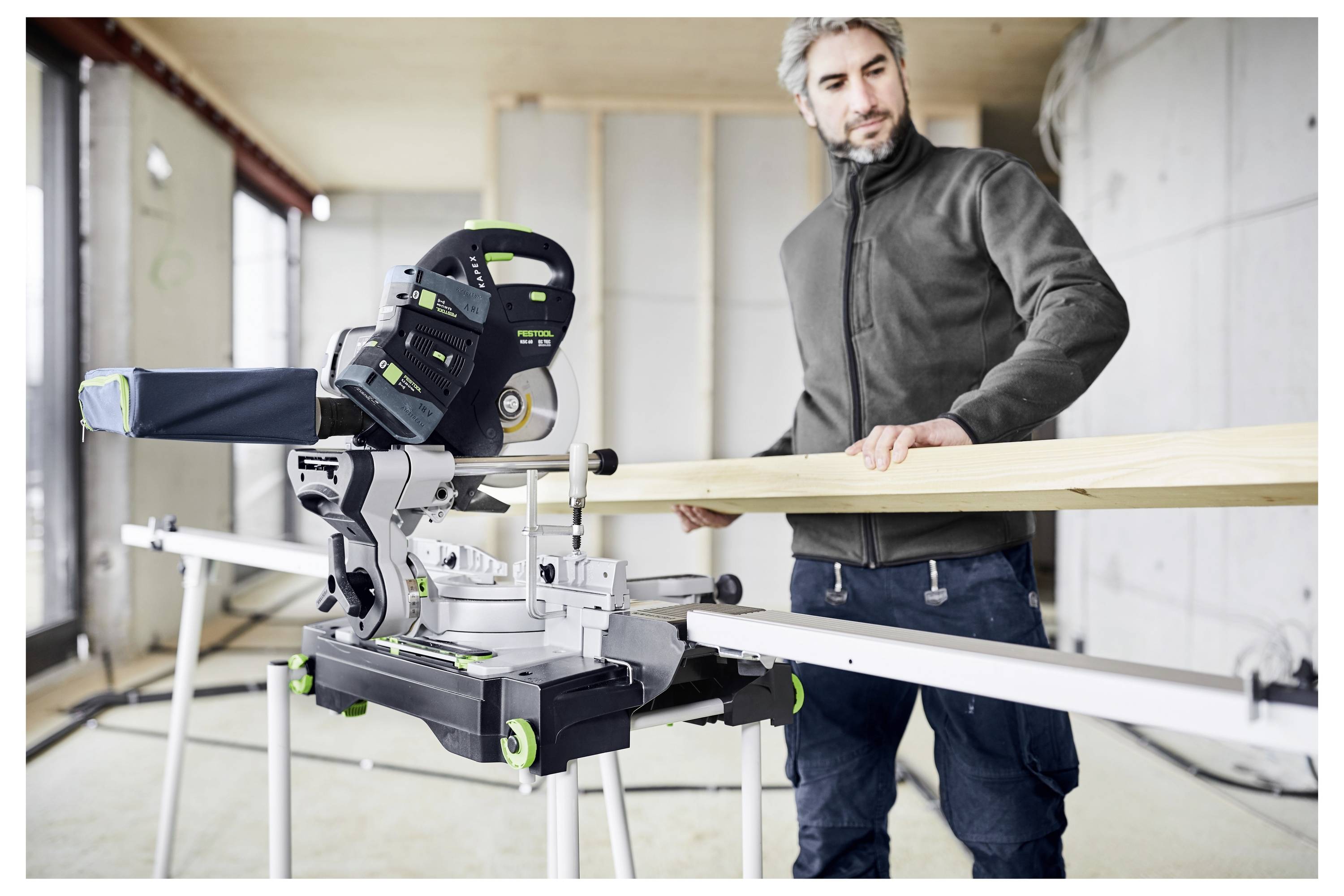 Una persona in un laboratorio utilizza una sega a squadra per tagliare una tavola di legno. L'ambiente è una stanza in costruzione parziale con pareti non finite.