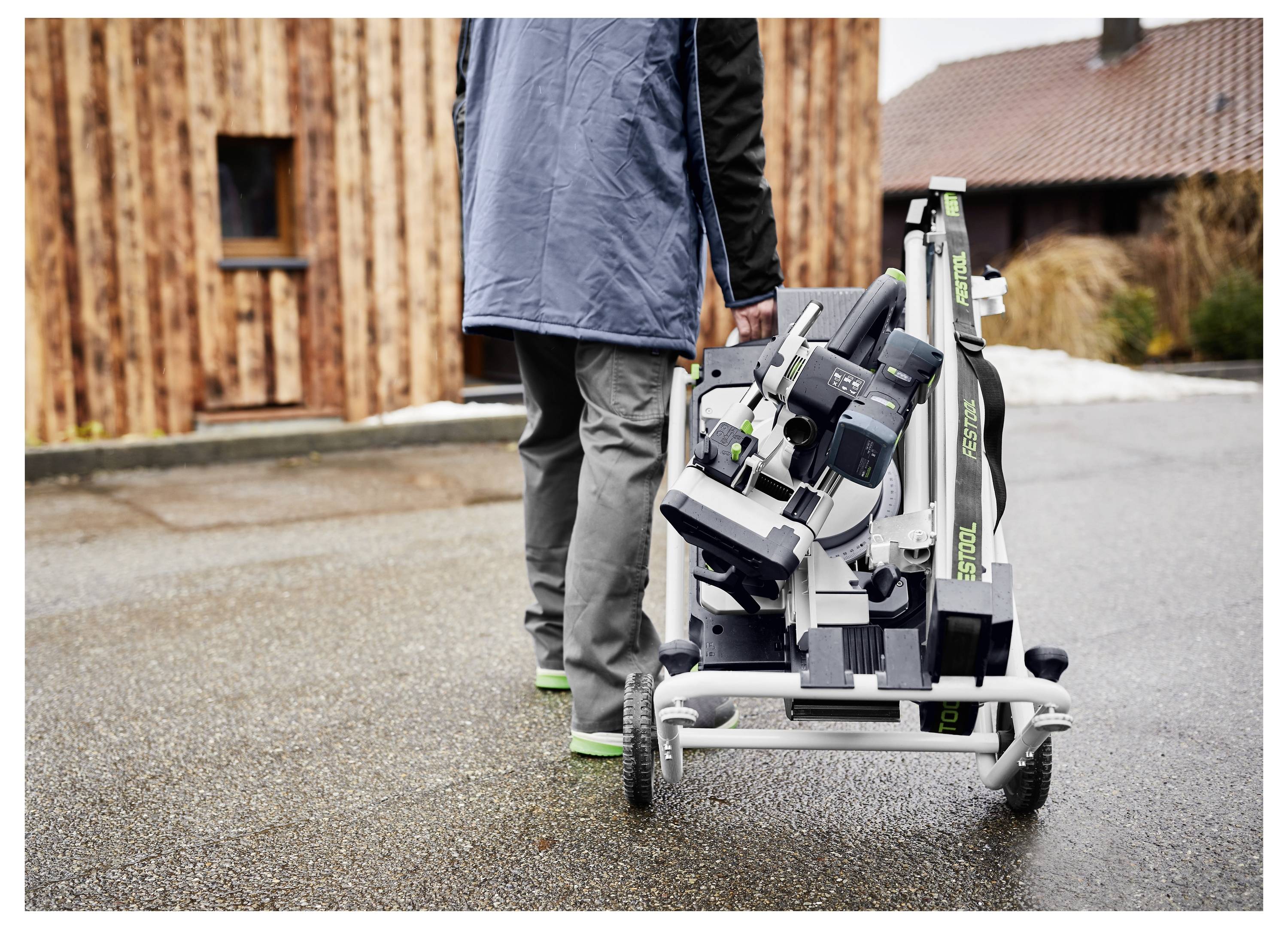 Una persona trasporta un'ascia pieghevole su un supporto portatile lungo un marciapiede bagnato, accanto a edifici in legno, il che implica la facile mobilità degli attrezzi.