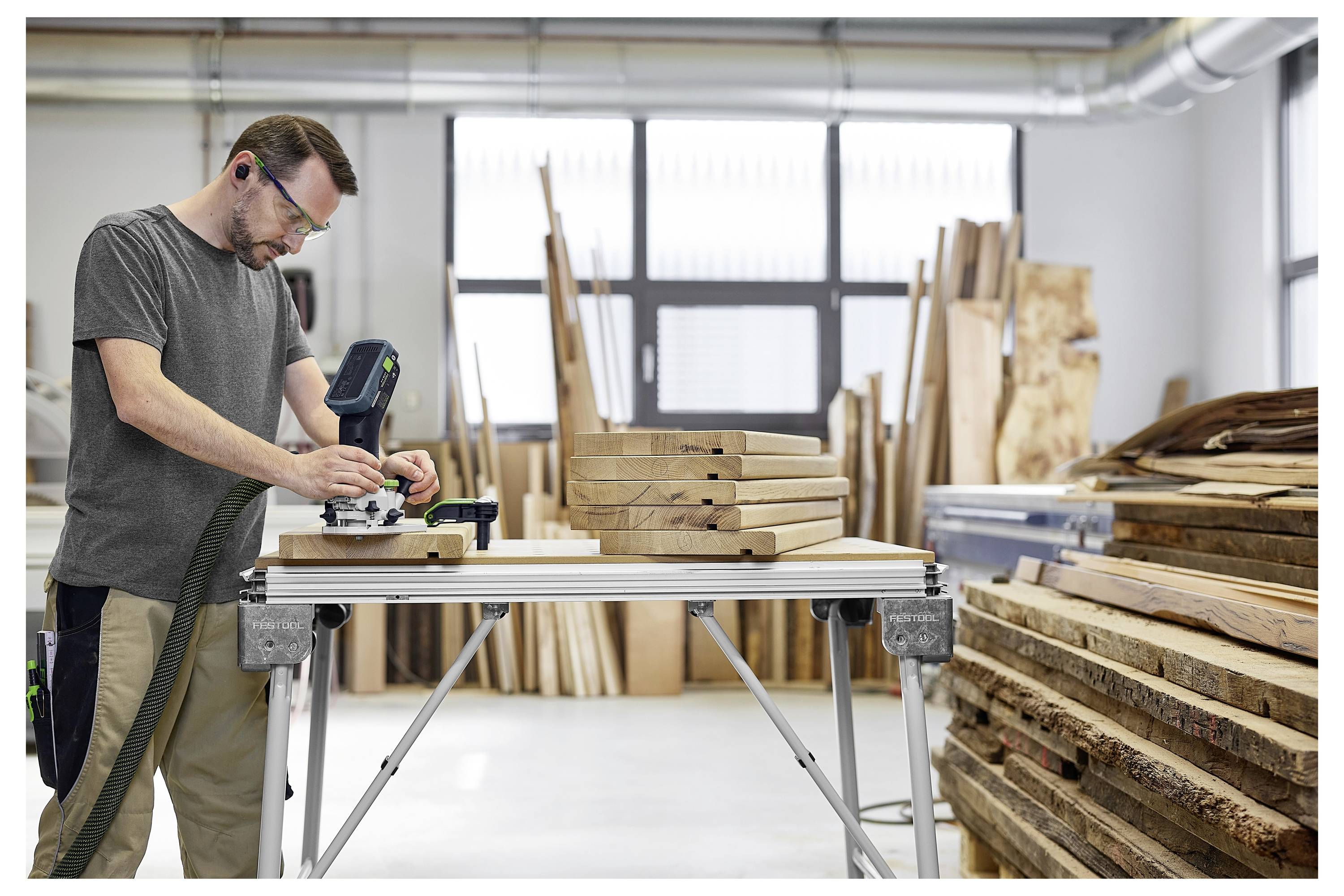 Un uomo che indossa occhiali di sicurezza utilizza un attrezzo elettrico su assi di legno in un laboratorio pieno di tavole di legno e luce solare proveniente da grandi finestre.