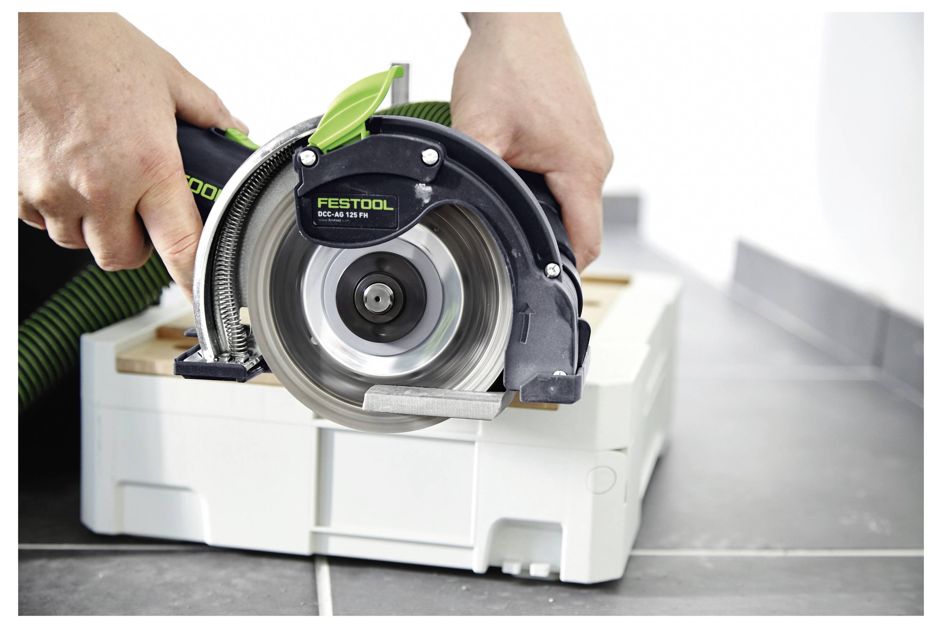 Una persona utilizza una sega circolare su una superficie in legno. La sega è nera e verde con una lama visibile. È collegata a un tubo di aspirazione.