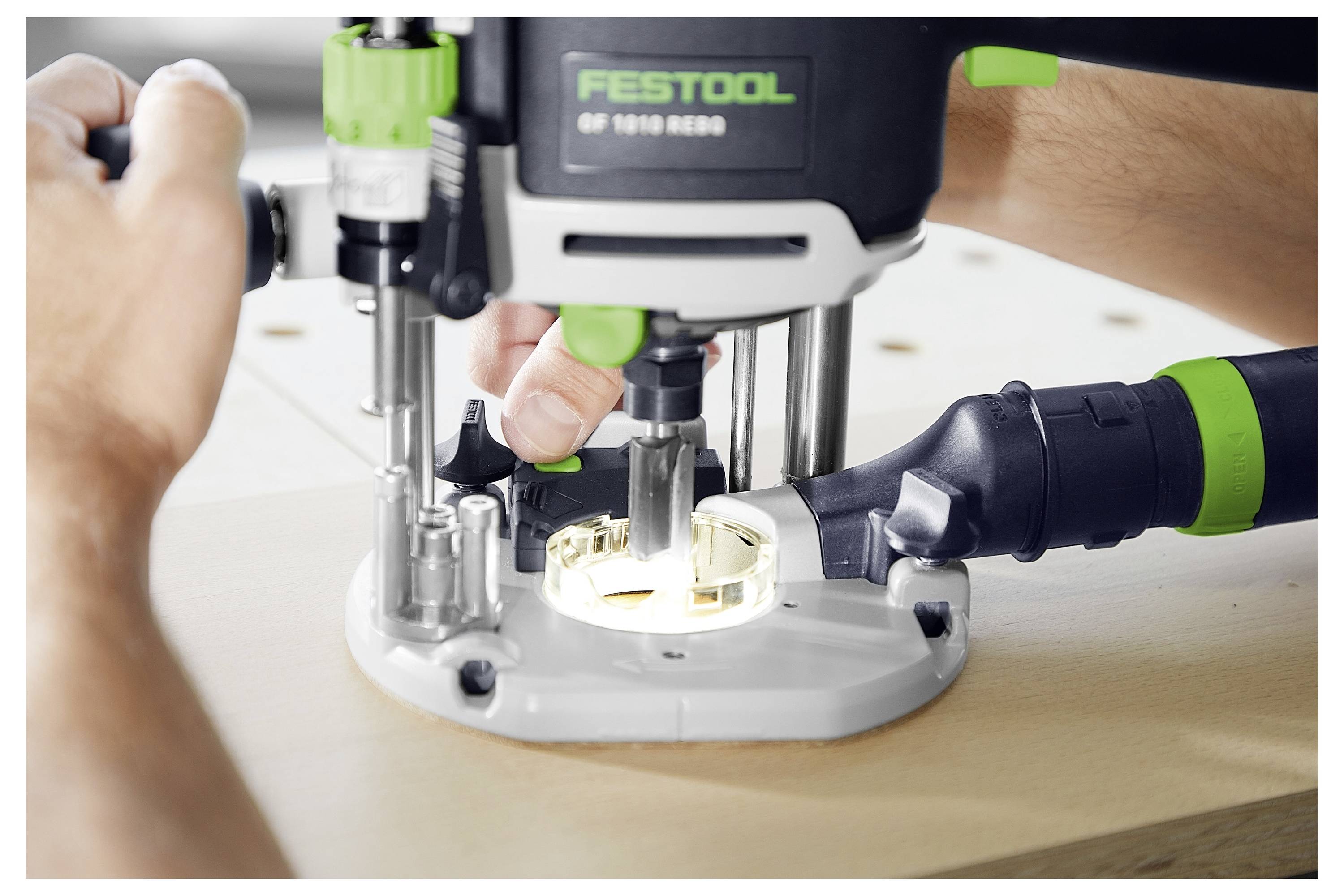 Una persona utilizza un router Festool su una superficie in legno, concentrandosi su un lavoro di precisione. Il router è nero e verde, lanciando trucioli di legno.