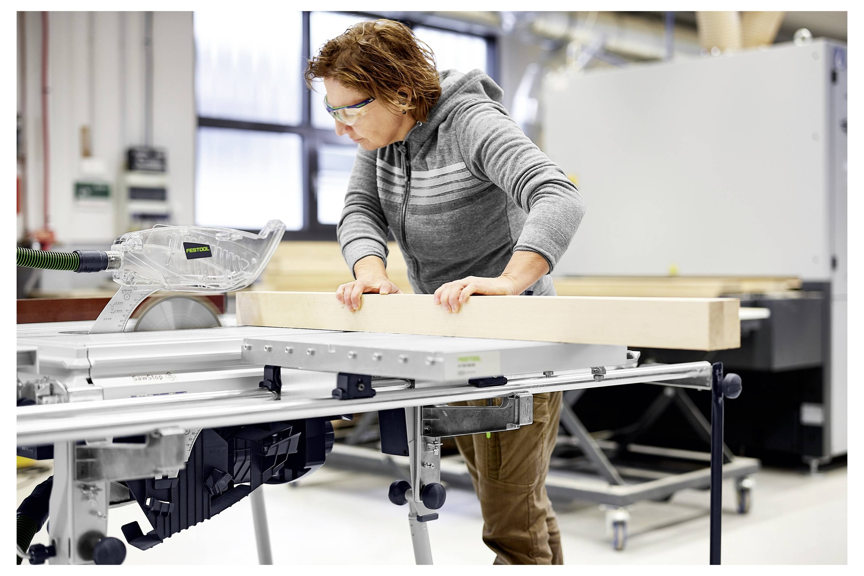 Una persona che indossa occhiali di sicurezza fa funzionare una sega circolare da tavolo, tagliando un grande pezzo di legno in un'officina.