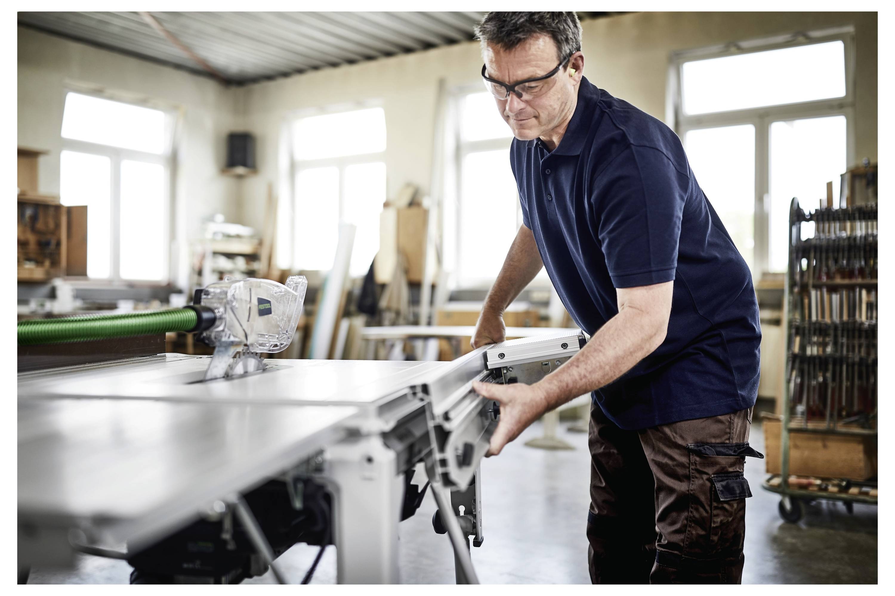 Un uomo con gli occhiali di sicurezza regola una sega circolare su tavolo in un laboratorio ben illuminato. Nel background sono visibili vari strumenti di falegnameria e materiali.