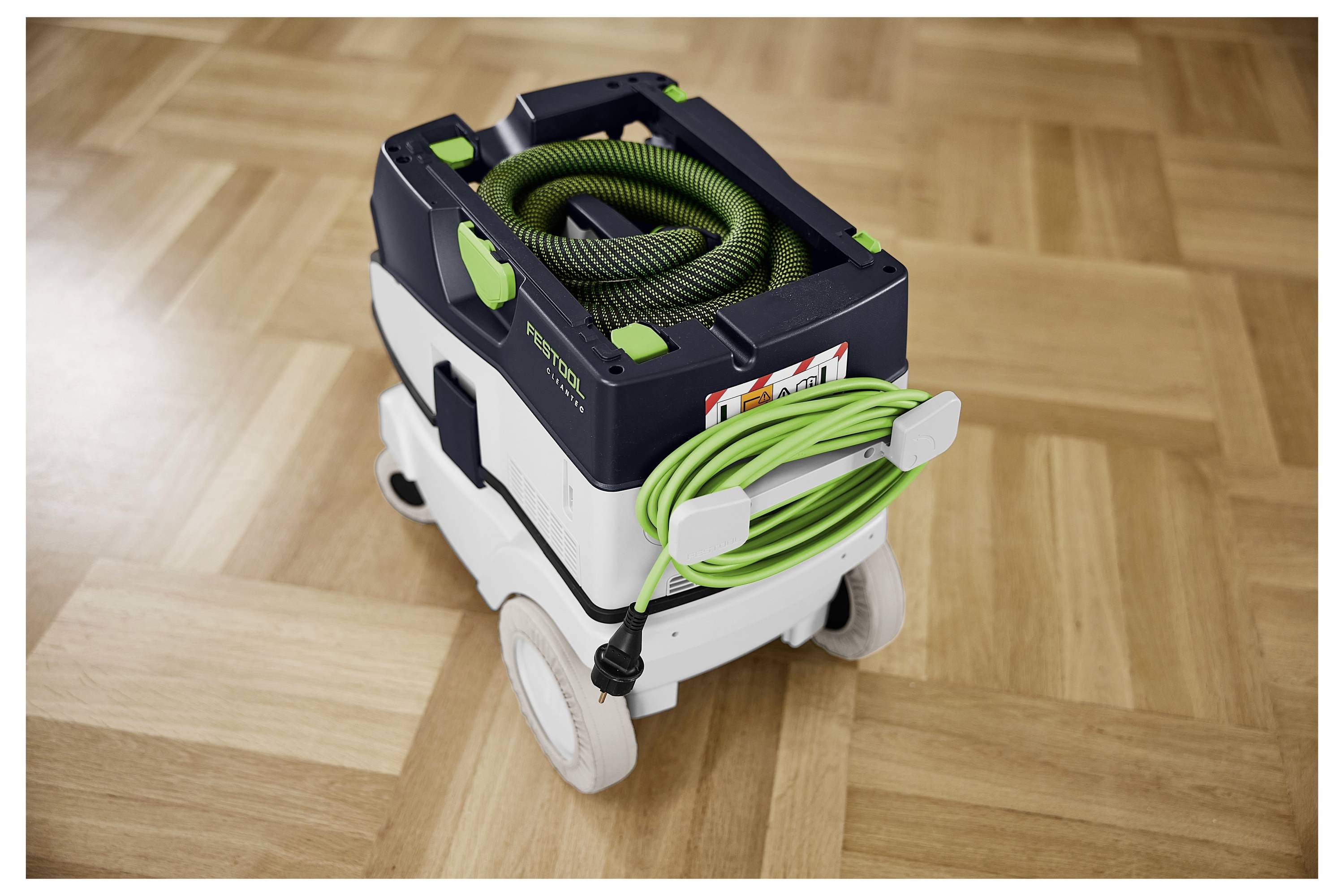 Un aspirapolvere portatile con un tubo verde arrotolato sulla parte superiore e sui lati, su un pavimento in legno, che mostra il suo design compatto.