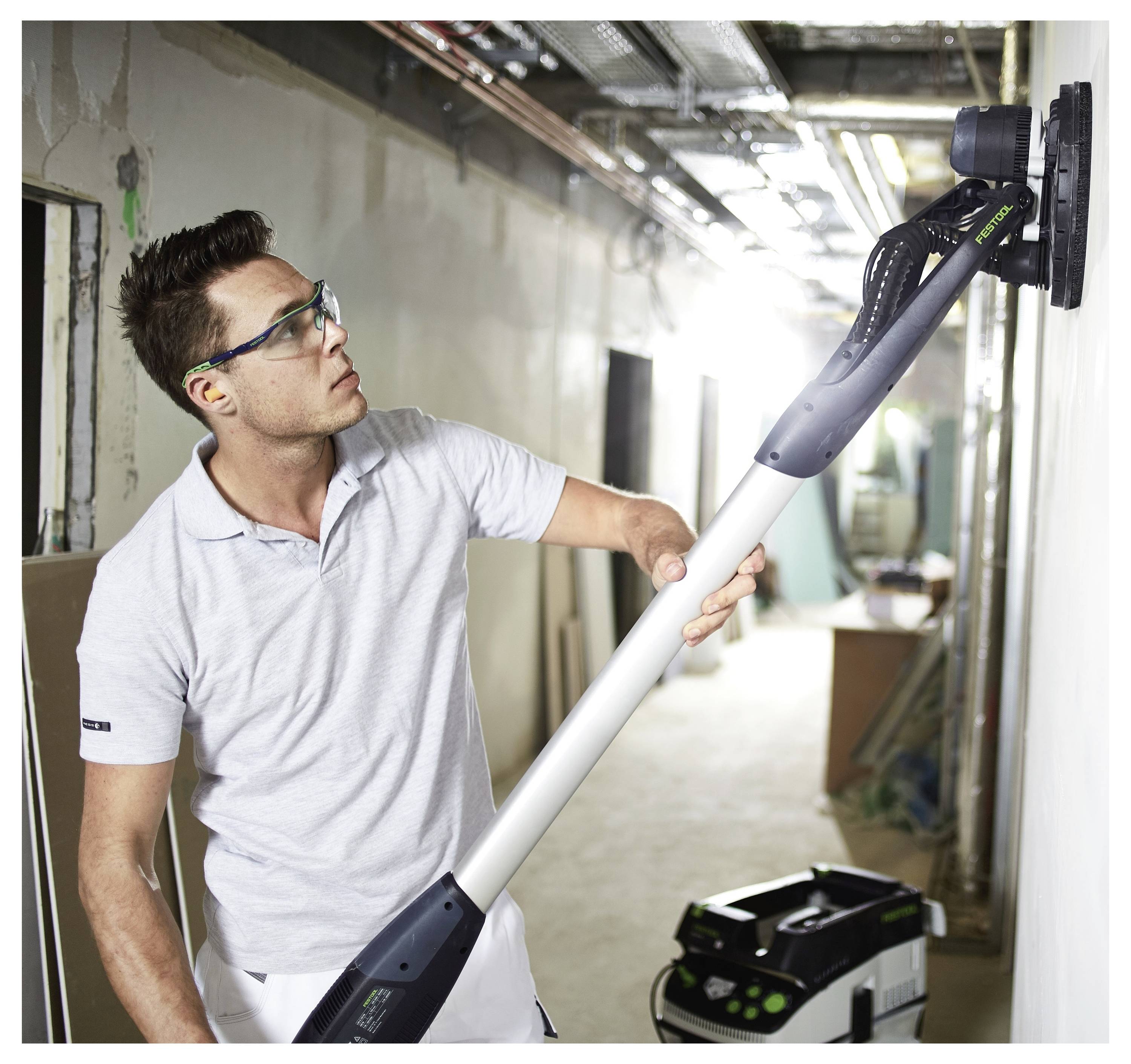 Una persona che indossa occhiali di sicurezza utilizza una levigatrice elettrica per levigare una parete in un'area di costruzione, con attrezzi e attrezzature nelle vicinanze.