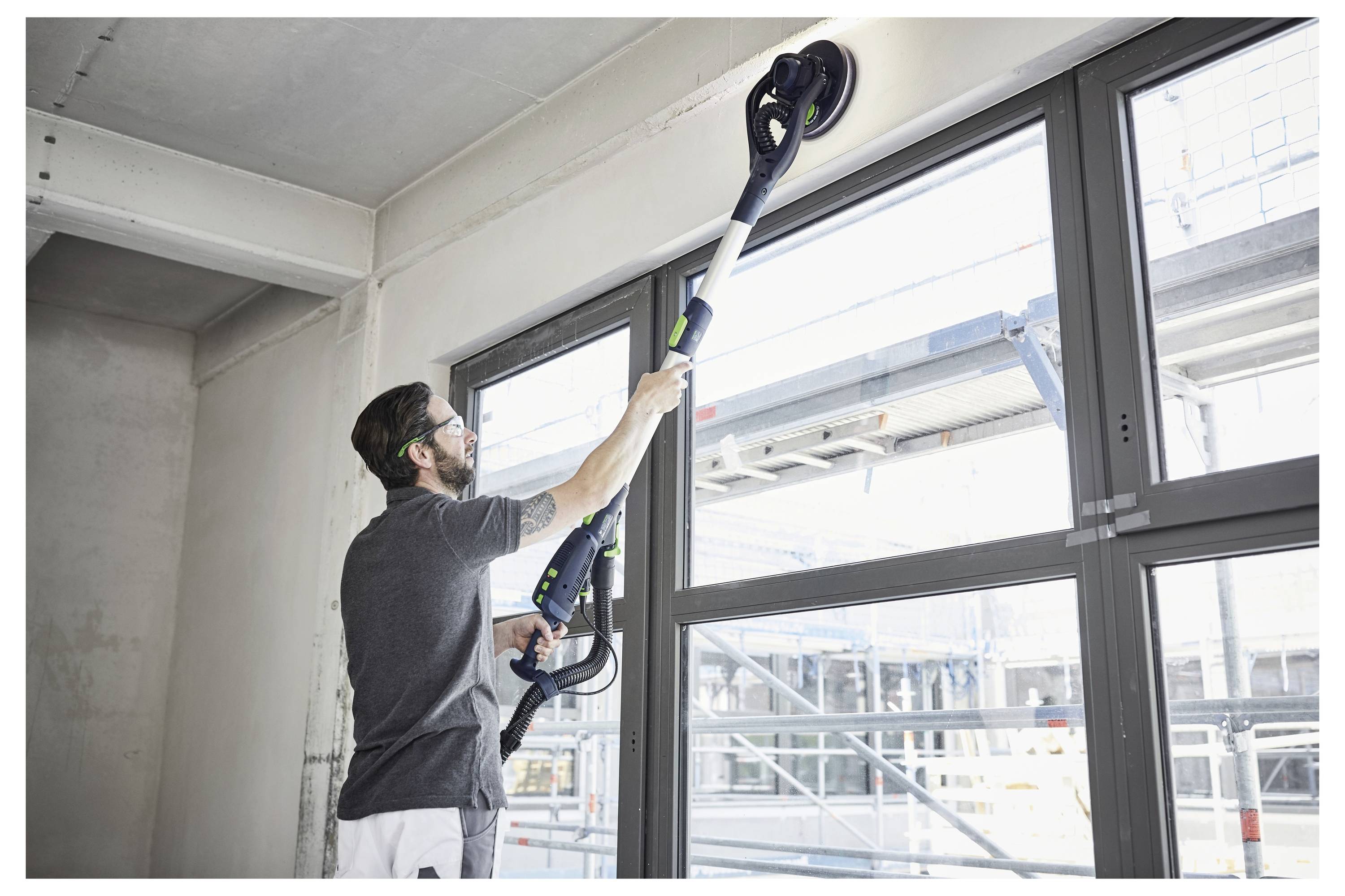 Persona che utilizza un utensile elettrico per carteggiare una parete alta vicino a grandi finestre in un edificio non completato, suggerendo lavori di costruzione o ristrutturazione.