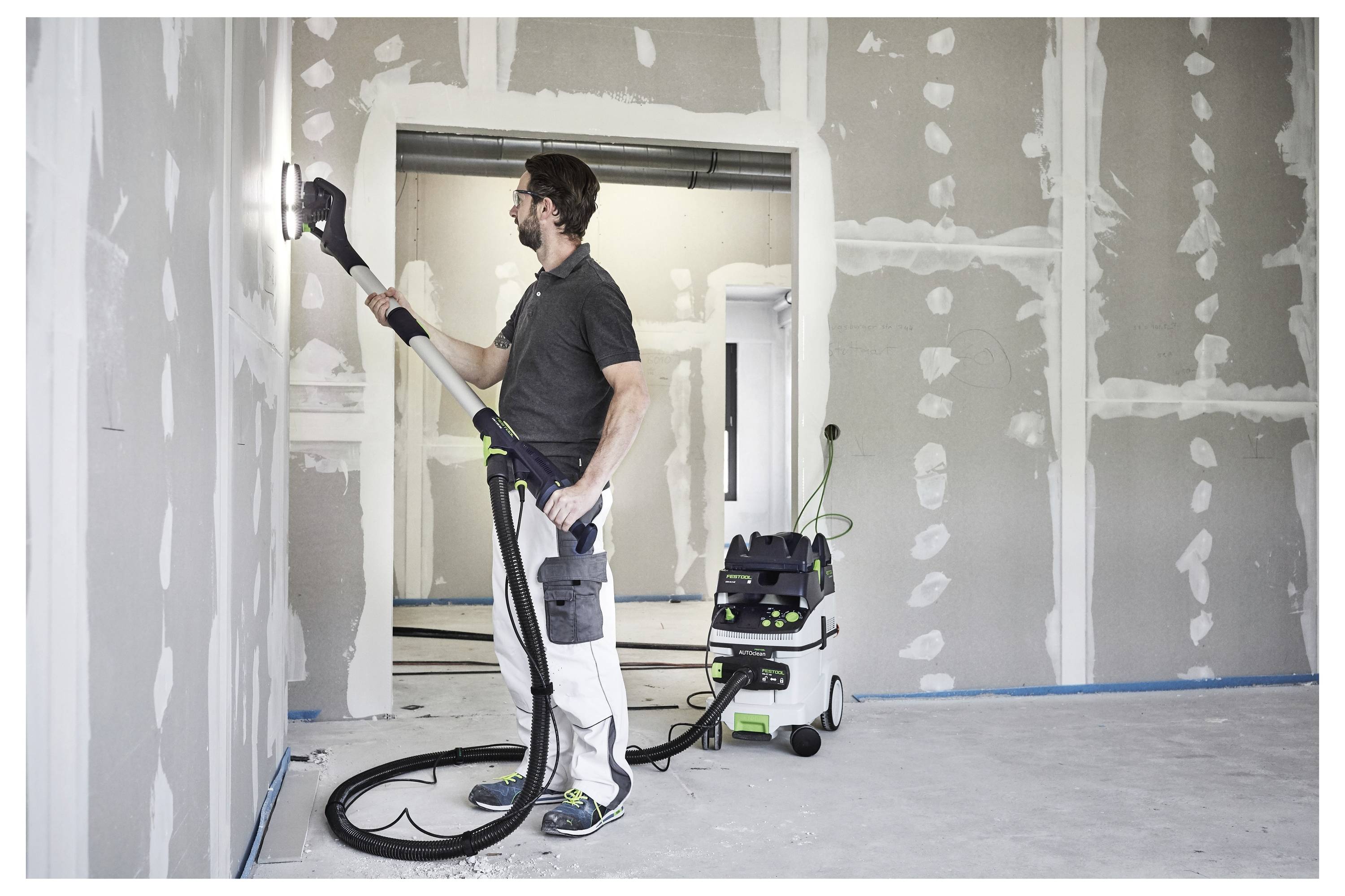 Un uomo leviga una parete in una stanza non finita utilizzando uno strumento di levigatura alimentato collegato a un sistema di aspirazione per raccogliere la polvere, promuovendo la pulizia.