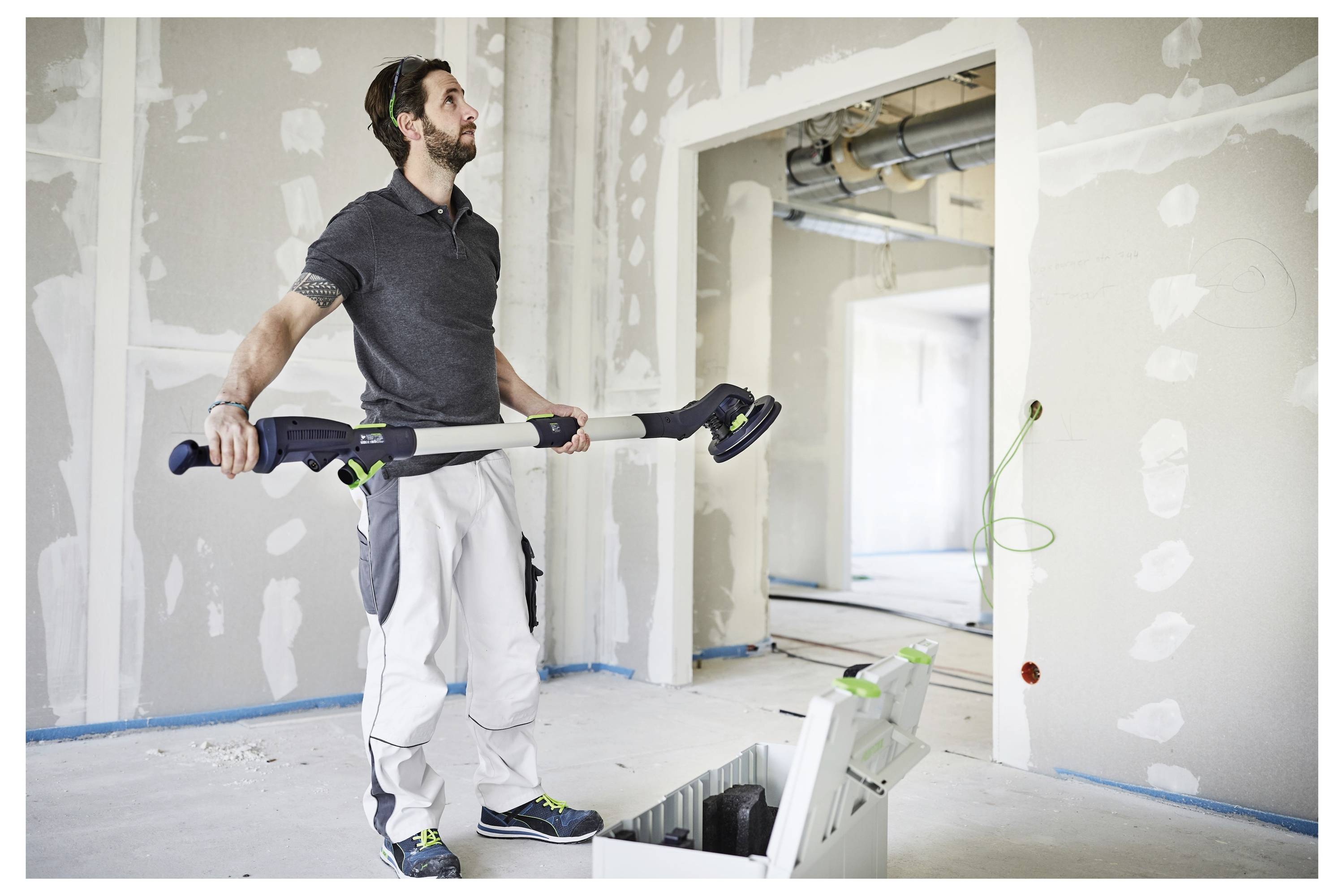 Una persona in una stanza parzialmente costruita tiene un grande attrezzo utilizzato per levigare le pareti, stando accanto a una cassetta degli attrezzi posata a terra.