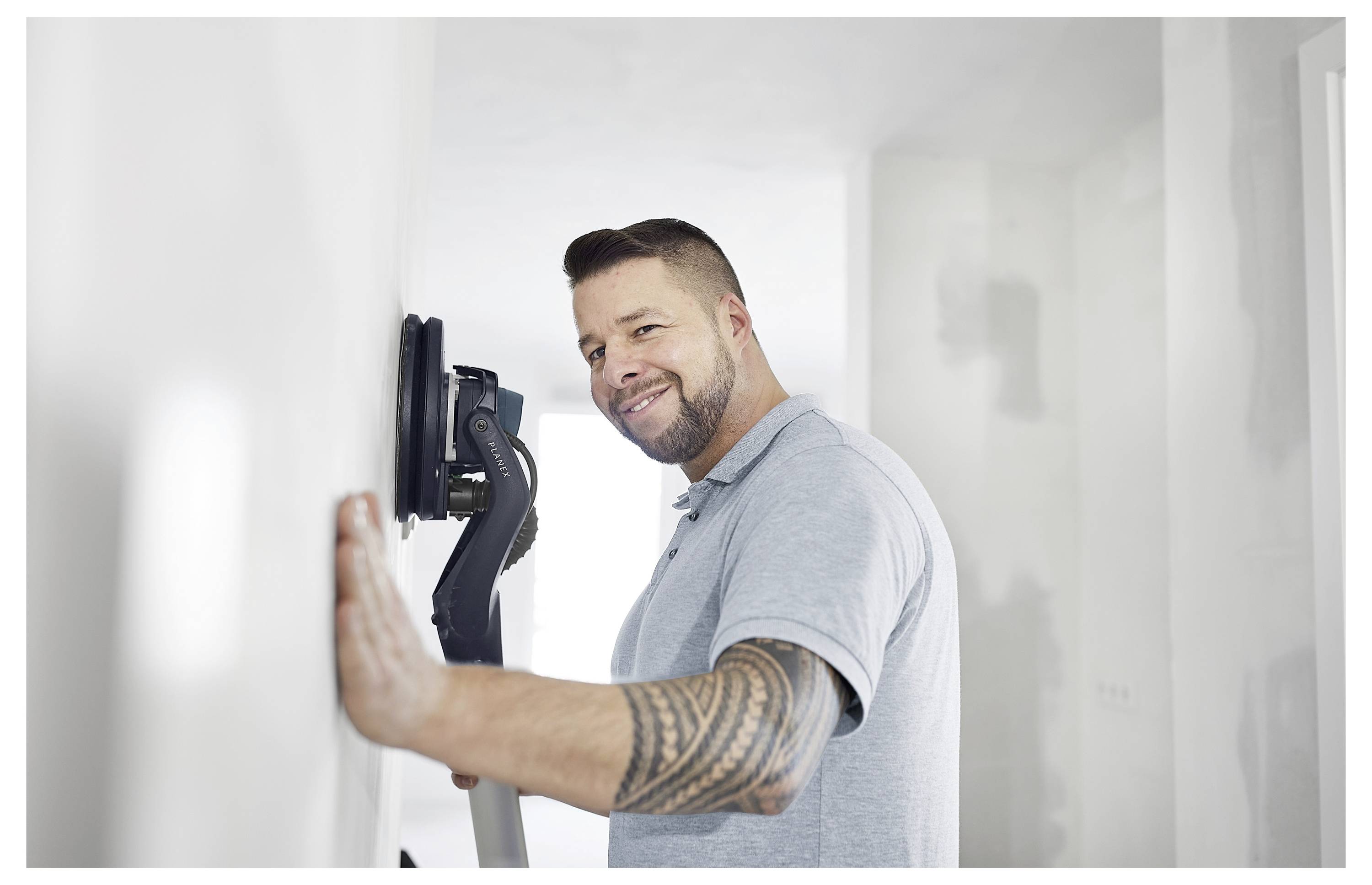 Uomo che utilizza una levigatrice a muro su una parete bianca, indossa una polo grigio chiaro, sorride, con un braccio tatuato, in una stanza parzialmente ristrutturata.