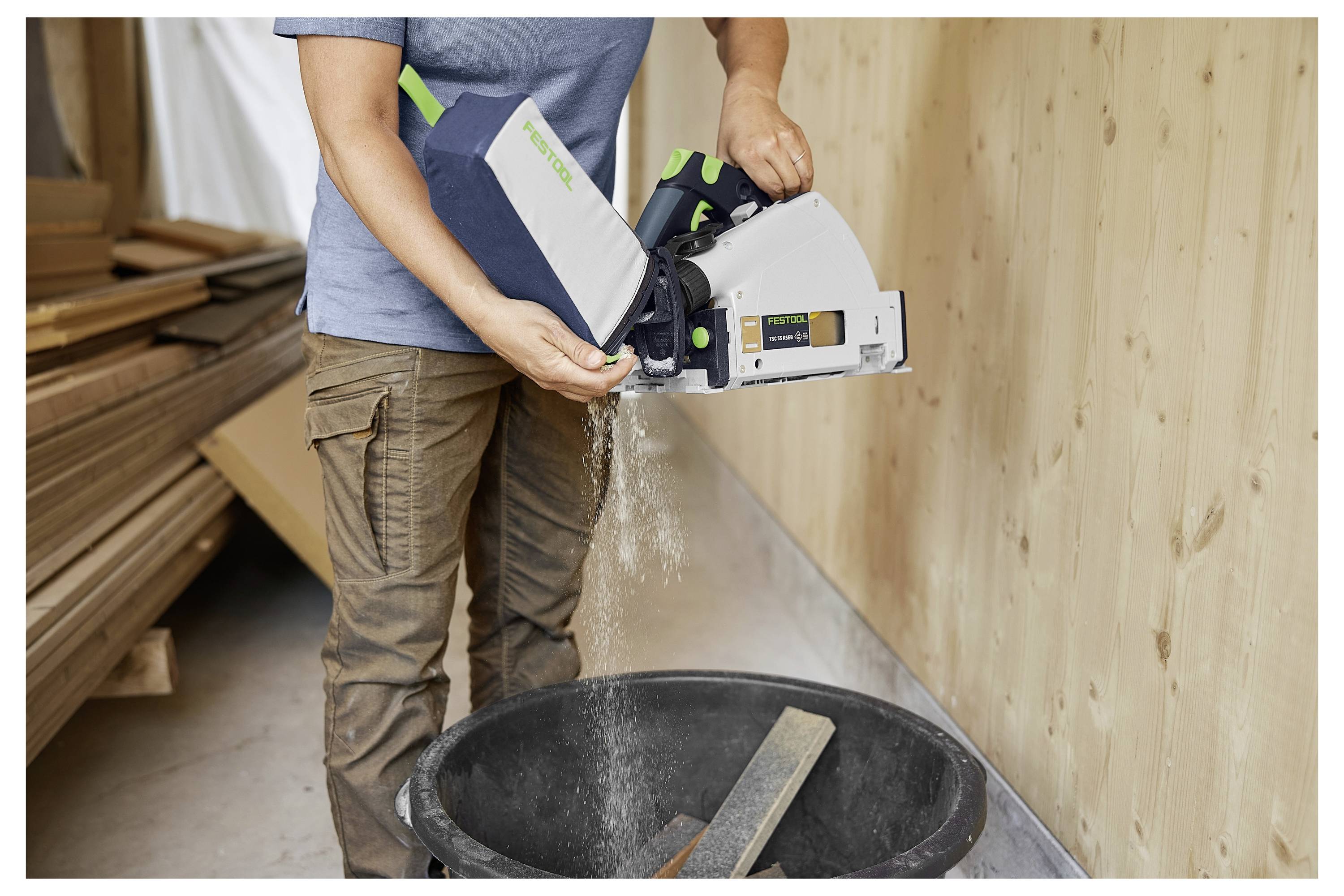 Una persona svuota i trucioli di legno da un aspiratore di polvere in un'officina. Nelle vicinanze, pile di legno appoggiano contro un muro.