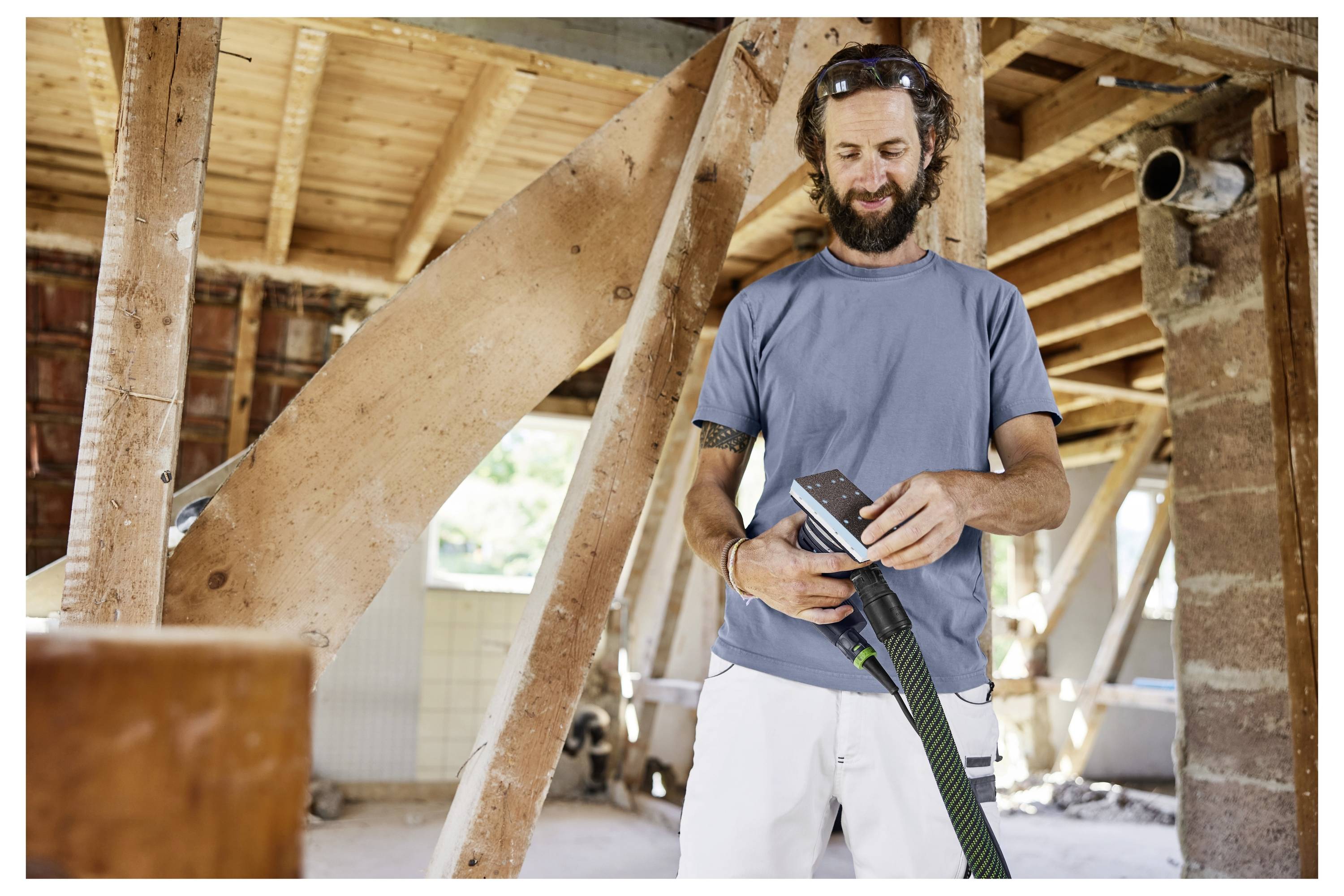 Un uomo in una camicia blu e pantaloni bianchi usa una levigatrice all'interno di un cantiere di costruzione in legno, sorridendo e concentrato sul suo lavoro.