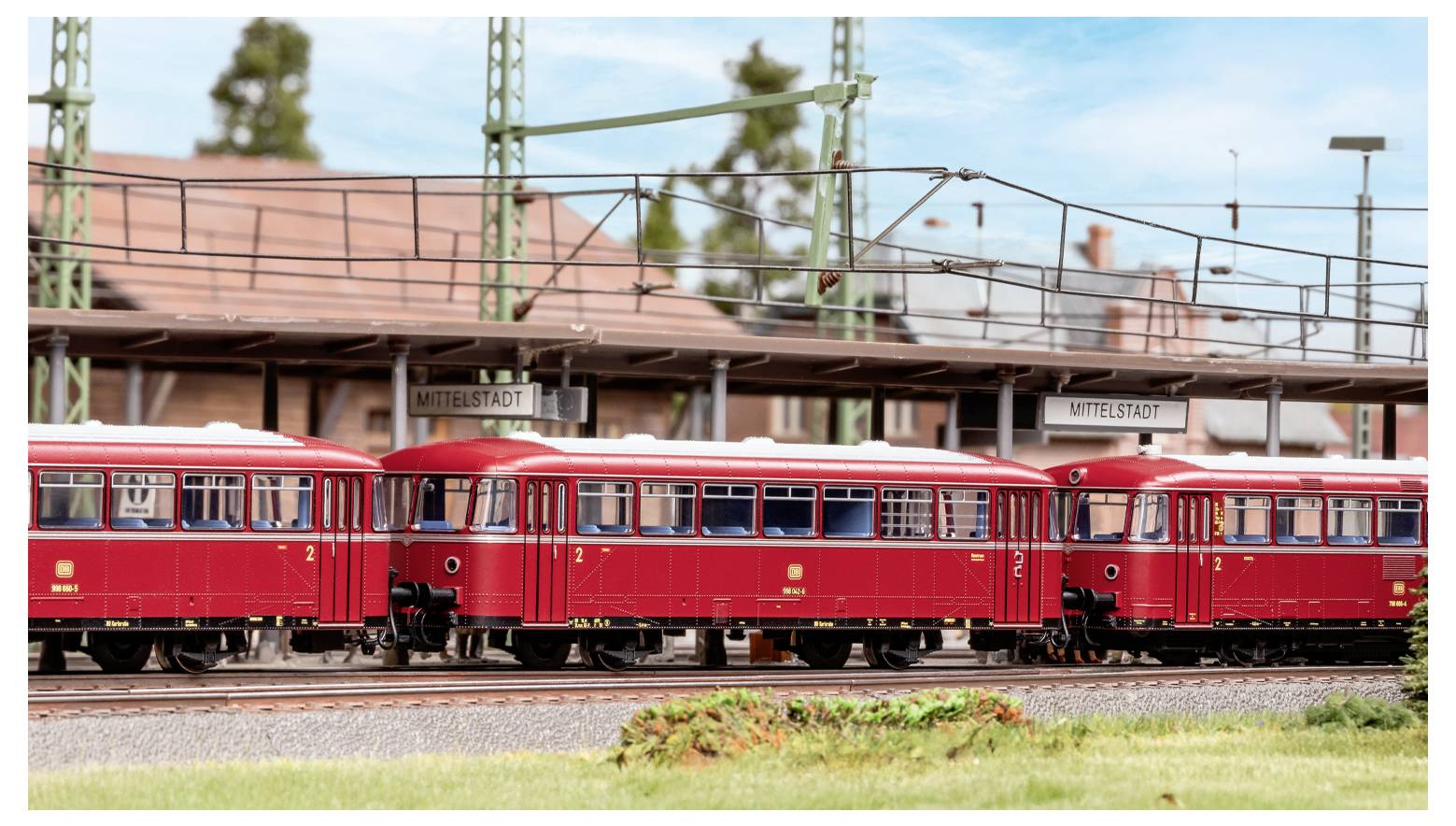 Una stazione ferroviaria modellistica con vagoni rossi etichettati 'Mittelstadt' sui binari. Presenta un'architettura dettagliata e uno scenario di sfondo elaborato.