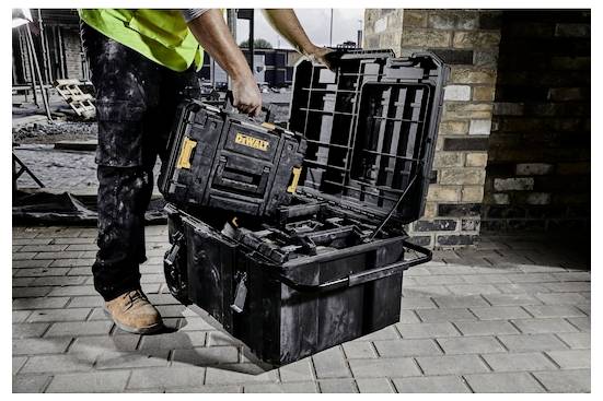 Una persona indossa stivali da lavoro e un giubbotto di sicurezza e posiziona una scatola di archiviazione degli attrezzi in una cassetta portautensili più grande su un cantiere con muri in laterizio visibili.