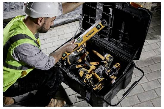 Un operaio edile in equipaggiamento di sicurezza recupera uno strumento a livella da una cassetta degli attrezzi piena di vari attrezzi da costruzione su una superficie pavimentata.