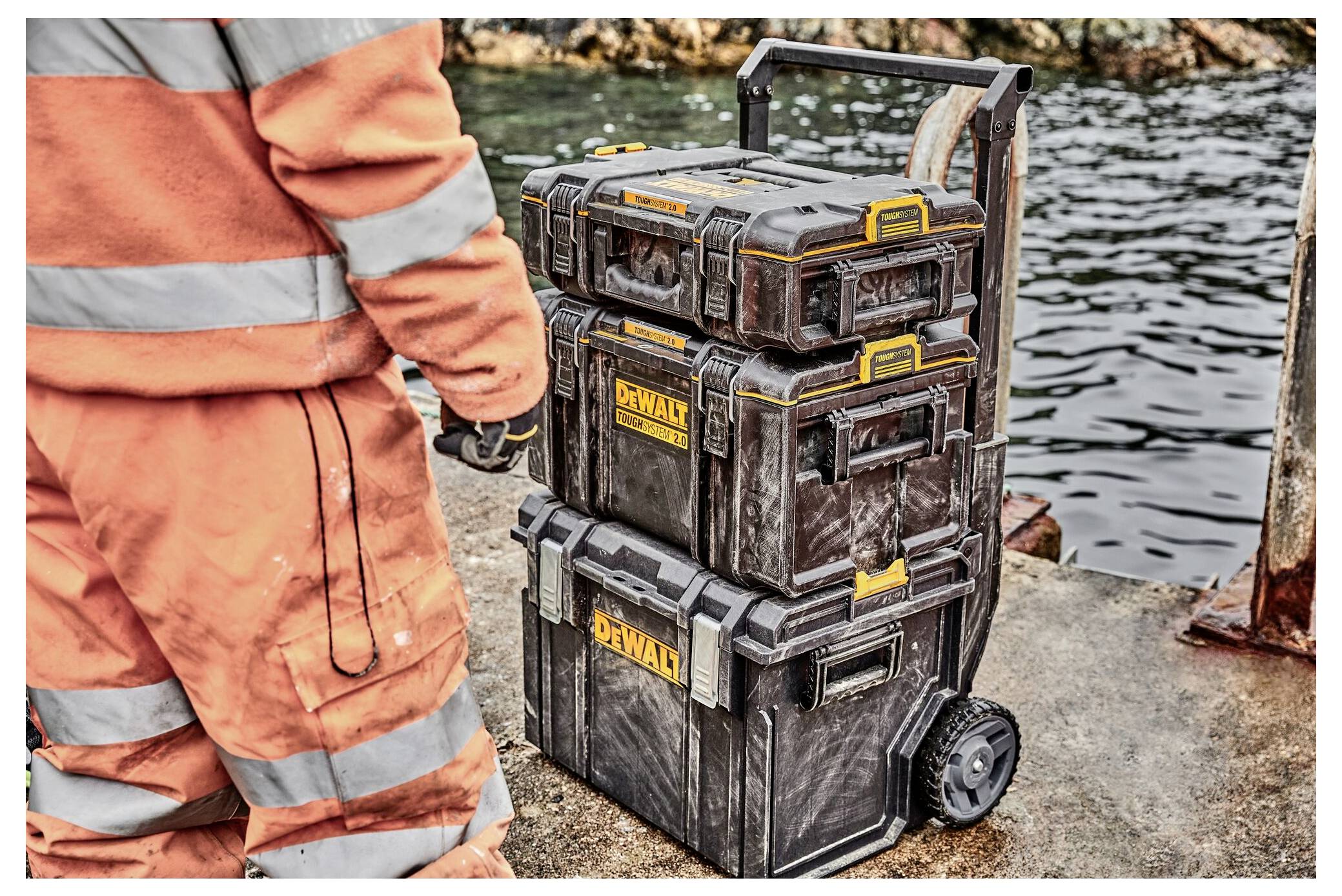 Un operaio in abbigliamento arancione si trova vicino a una pila di cassette degli attrezzi DeWalt accanto all'acqua su una superficie in cemento. L'allestimento è per uso industriale.