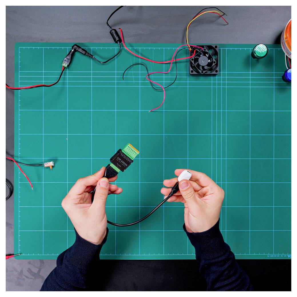 Mani che tengono connettori elettronici su un tappetino di lavoro verde con fili, una ventola e piccoli componenti, che suggerisce l'assemblaggio di componenti elettronici.