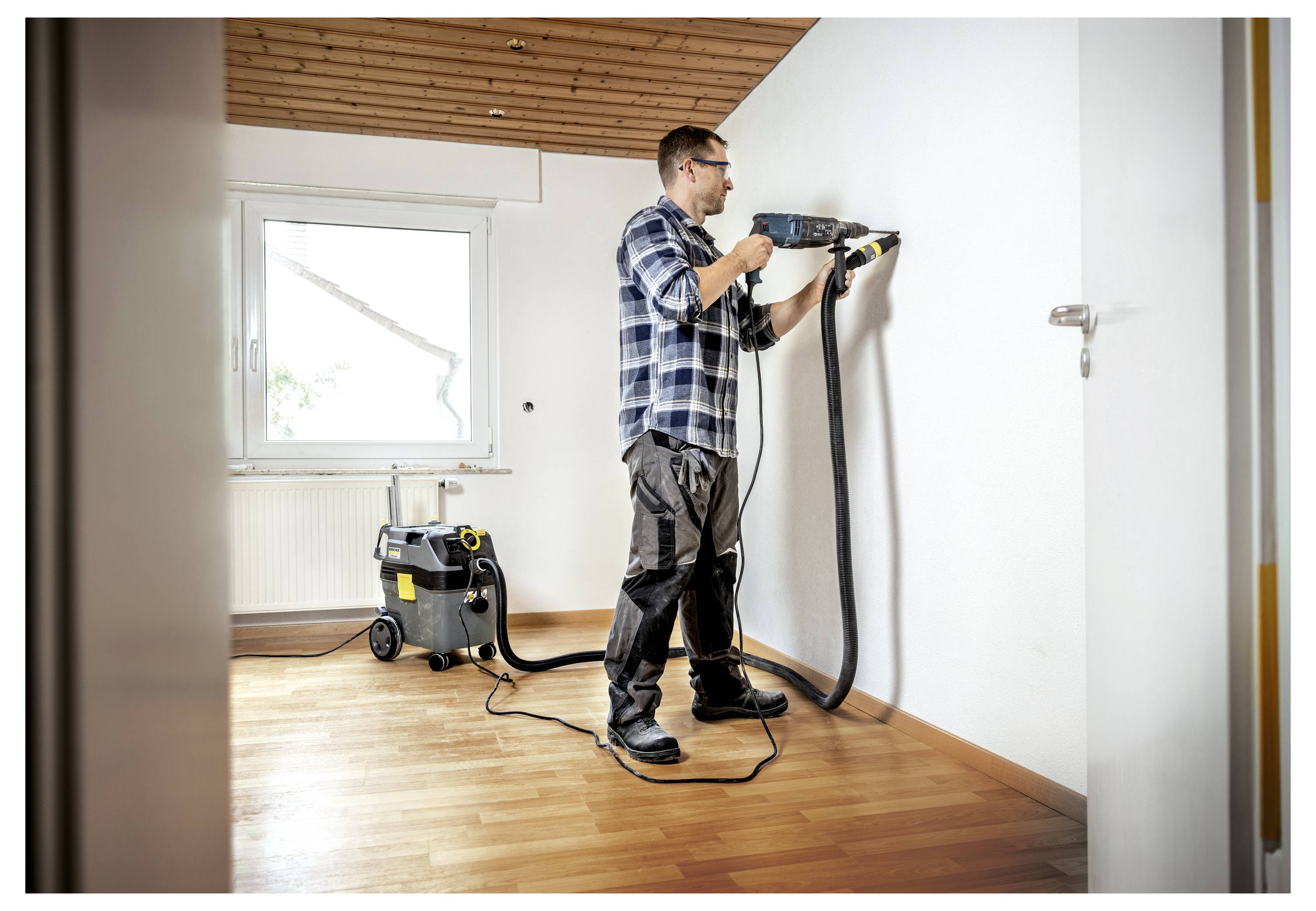 Una persona vestita in abbigliamento da lavoro casual usa un attrezzo elettrico collegato a un aspirapolvere per carteggiare o forare la parete in una stanza luminosa e parzialmente arredata.