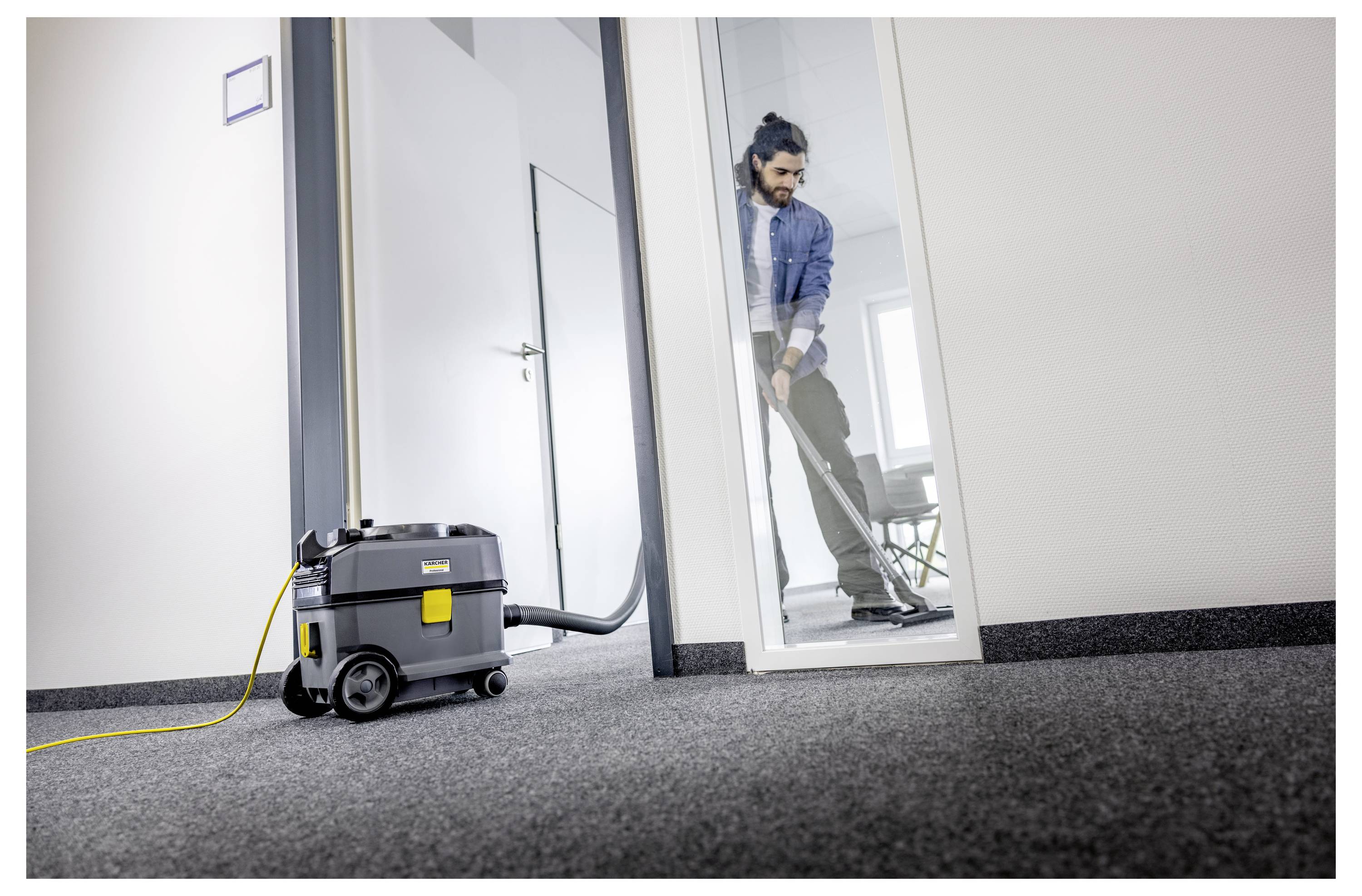 Una persona che aspira un tappeto in un ufficio usando un aspirapolvere grigio, con una porta in vetro visibile sullo sfondo.