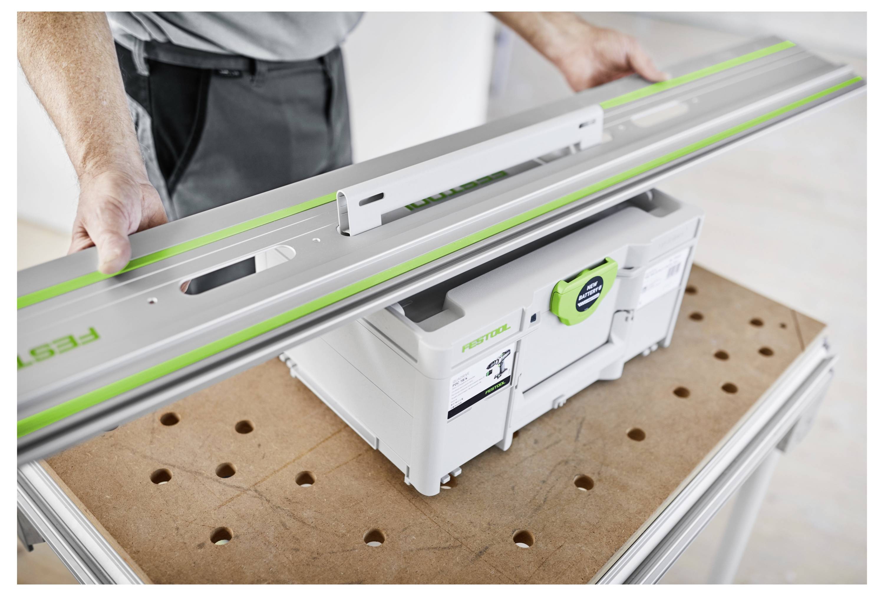 Persona che tiene un lungo e piatto utensile elettrico sopra una cassetta di immagazzinamento Festool verde e bianca su un banco da lavoro perforato.