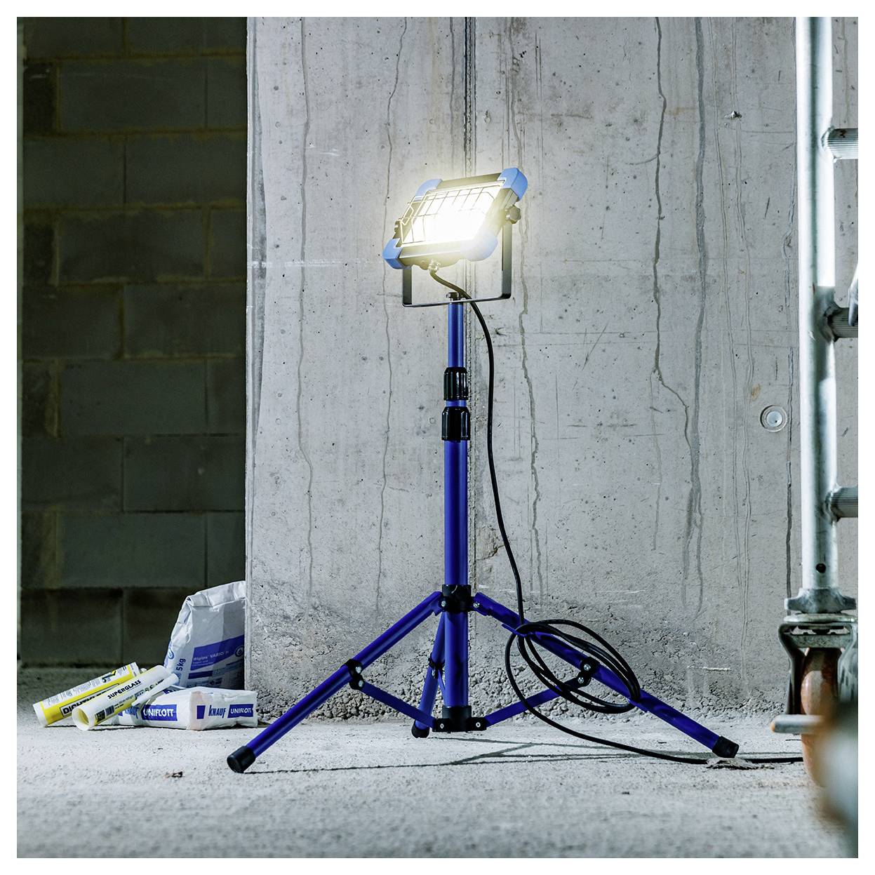 Una luce da lavoro luminosa su un treppiede blu si trova su un pavimento di cemento, illuminando un cantiere con muri in cemento e materiali da costruzione.