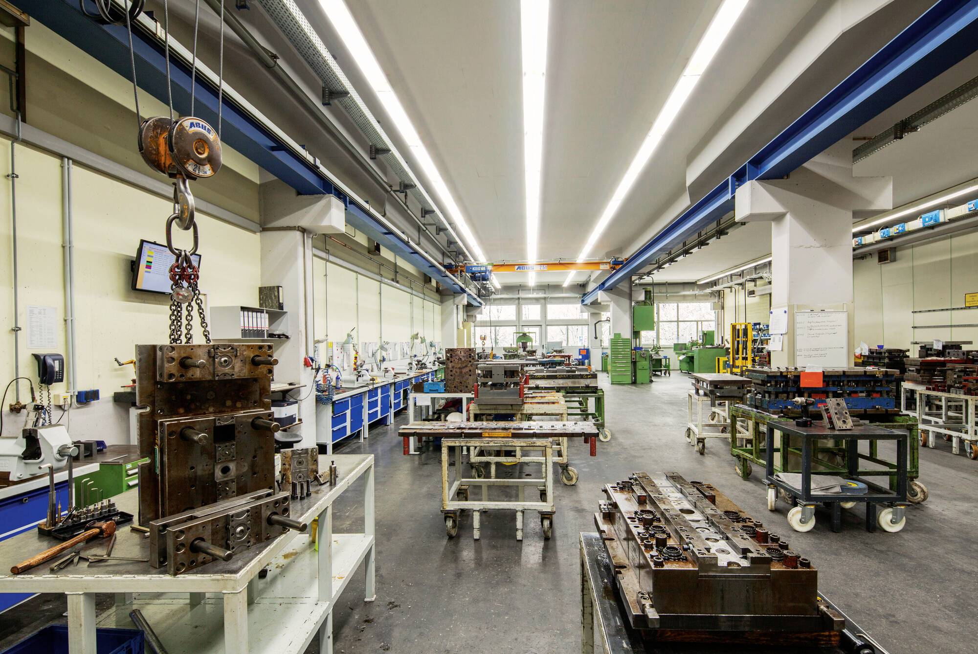 Interno di officina metallurgica con macchinari, banchi di lavoro e gru aerea. Attrezzi e attrezzature sono organizzati lungo le pareti e i tavoli.
