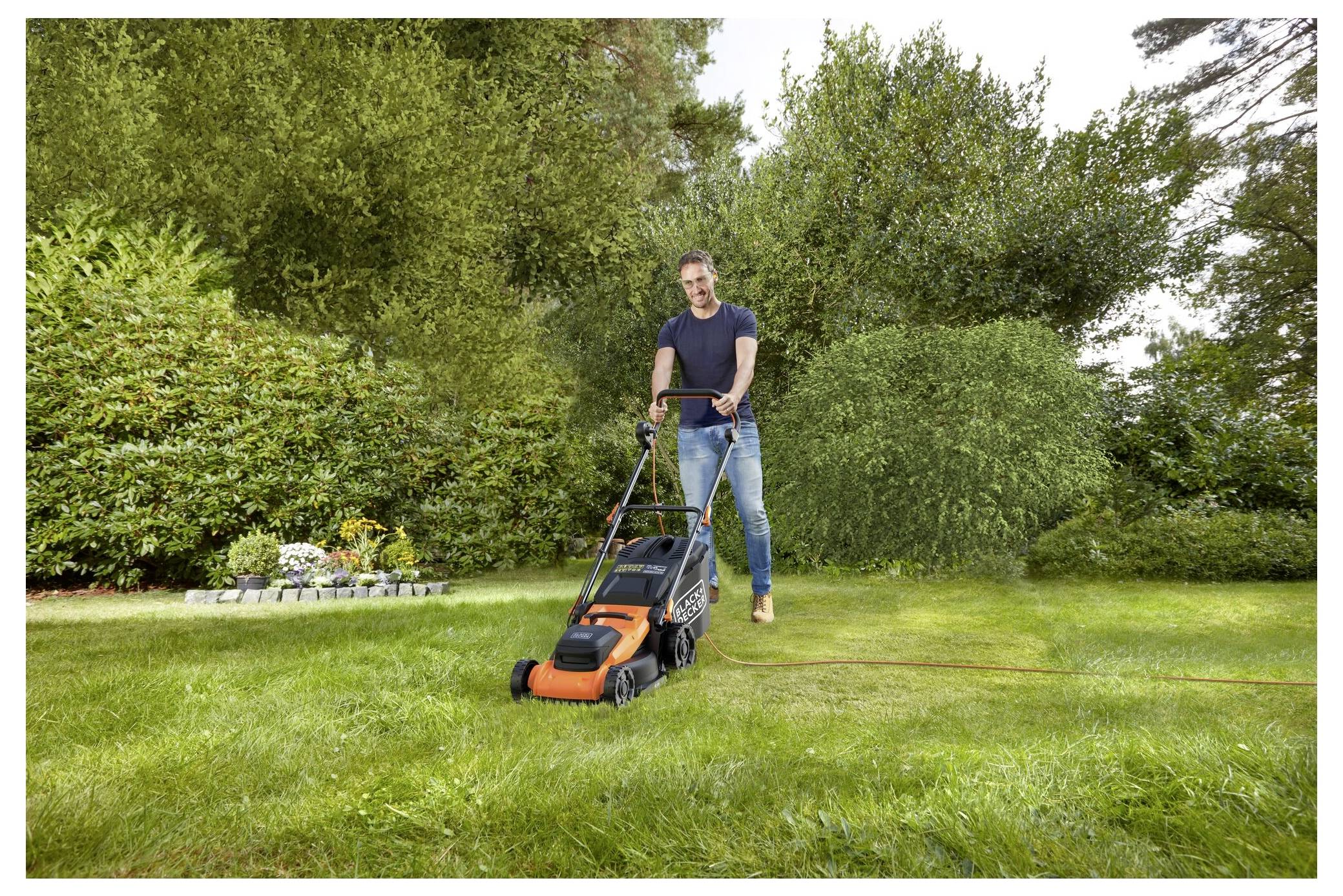 Una persona taglia un prato lussureggiante e verde con un tosaerba arancione e nero. Alberi e arbusti circondano il giardino sullo sfondo.