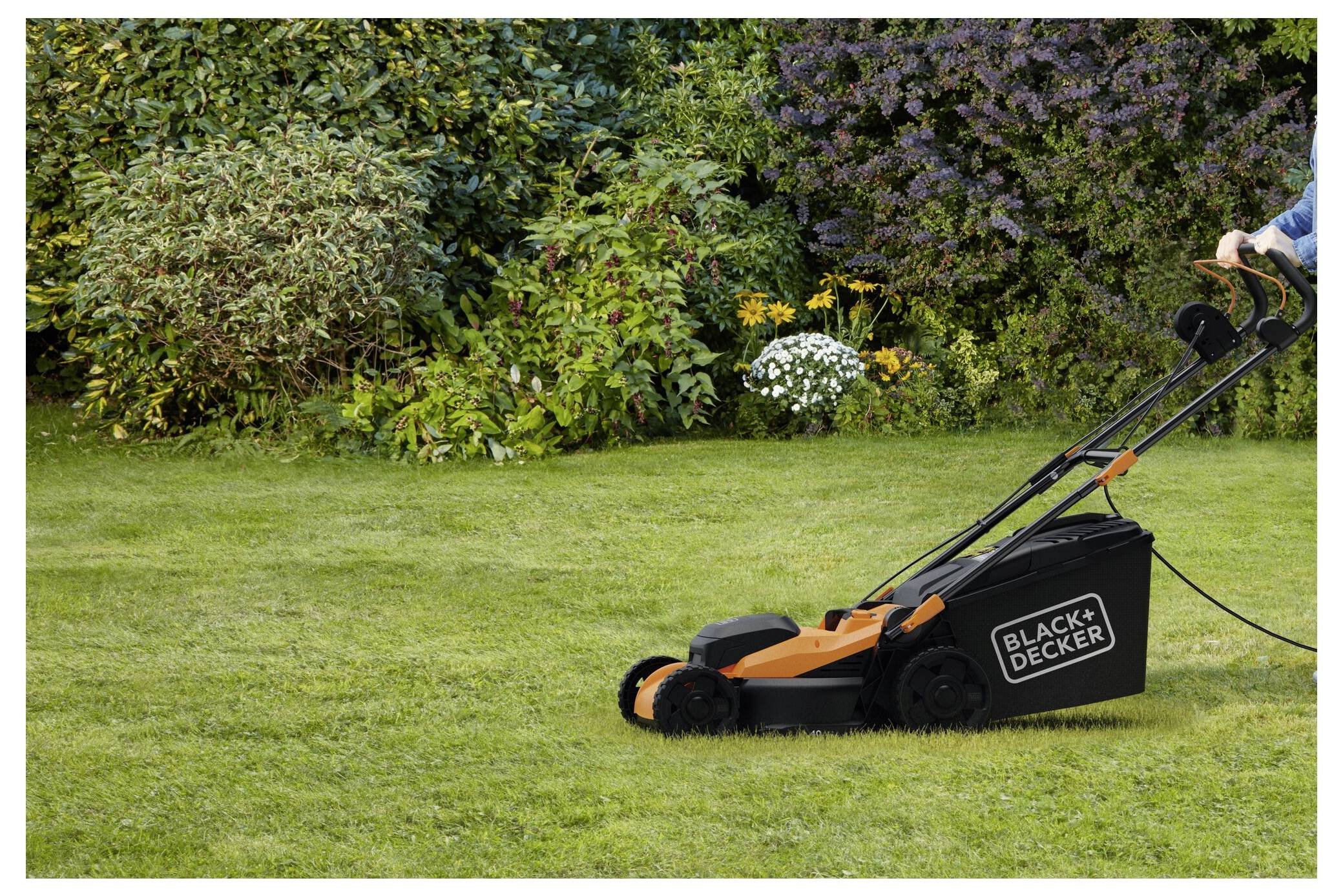 Una persona sta usando un tosaerba Black+Decker su un prato ben curato, circondato da cespugli lussureggianti e verdi e piante fiorite.