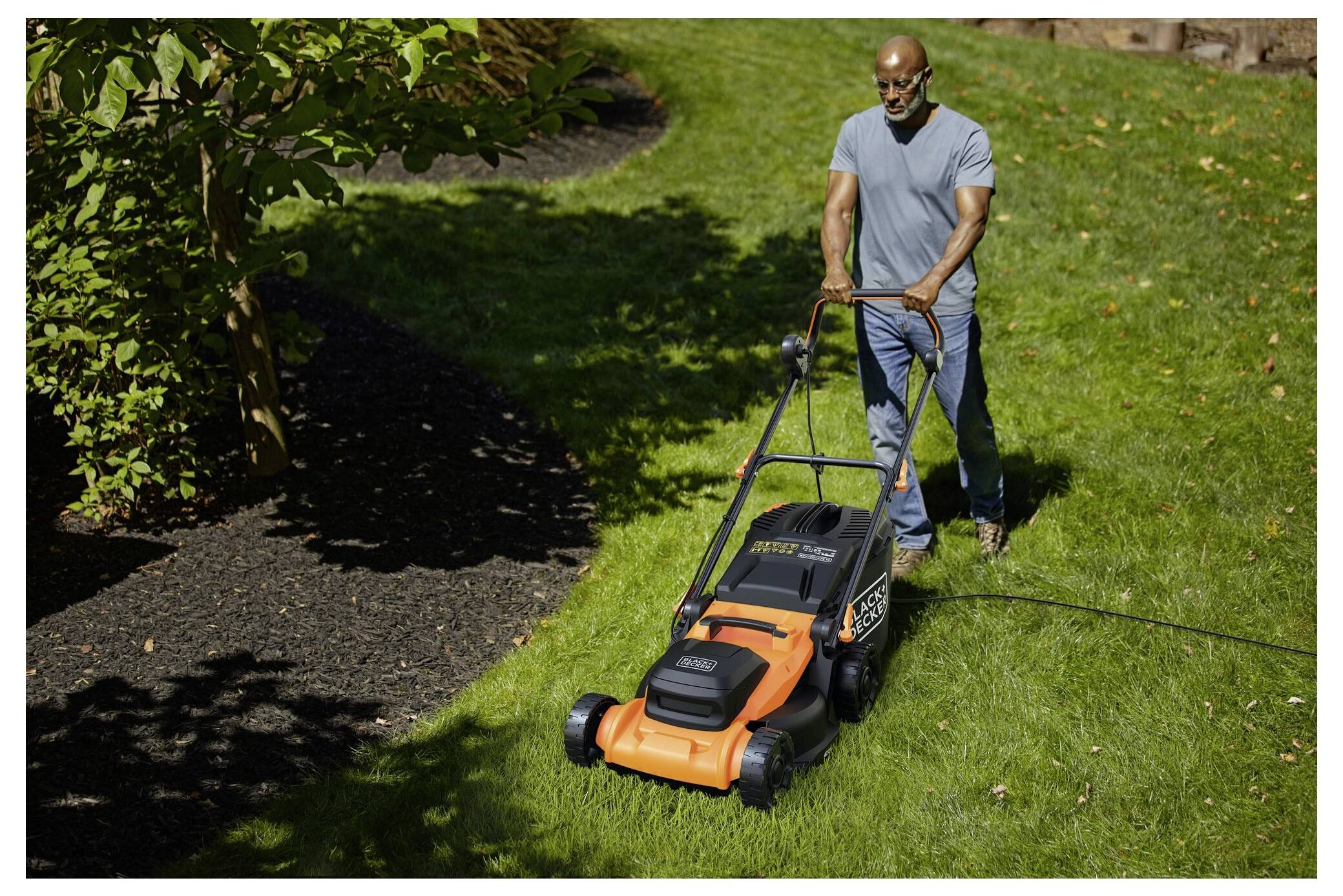 Una persona che taglia un prato verde e vibrante con un tosaerba elettrico arancione e nero, circondata da alberi e arbusti in ombra.