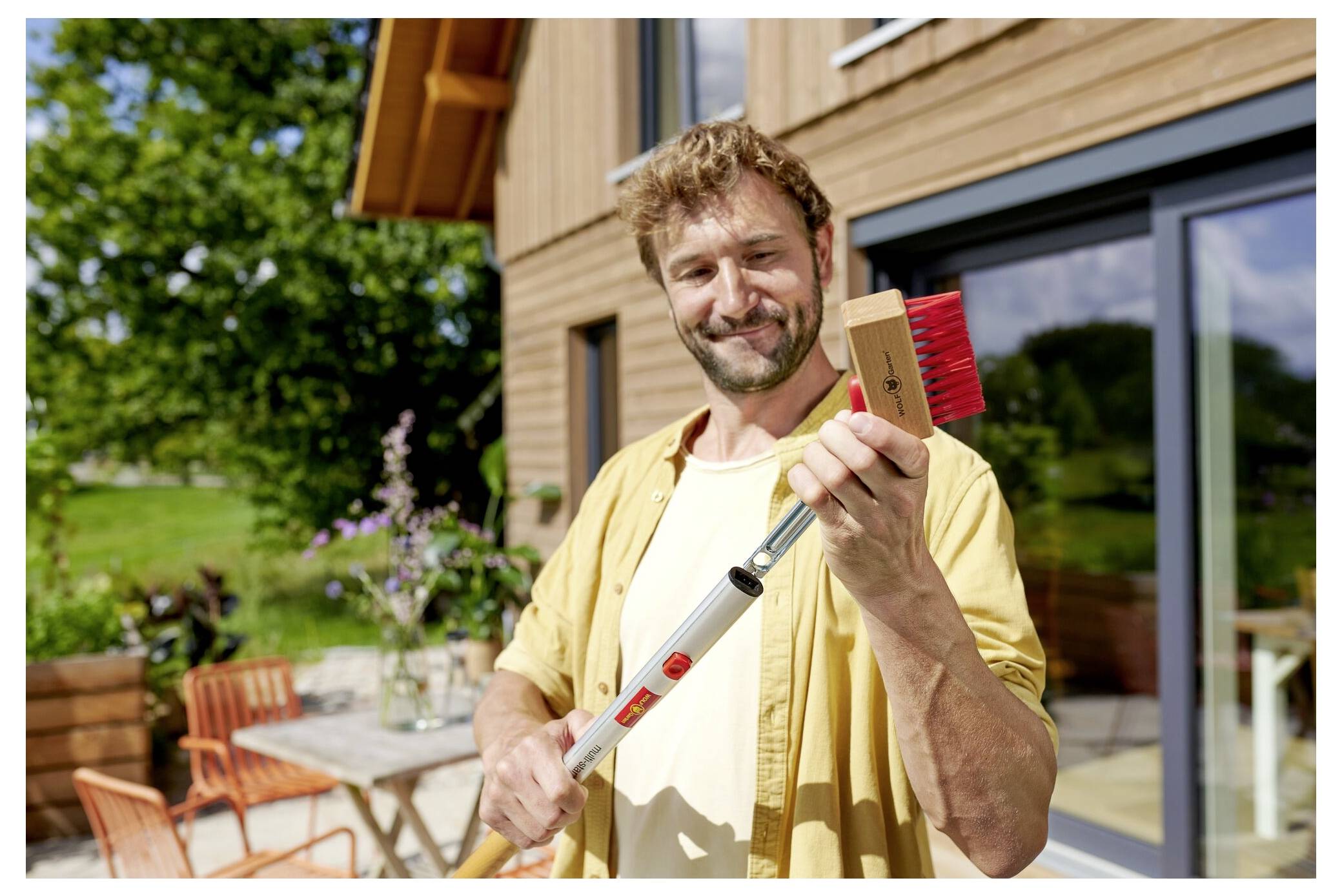 Una persona che sorride e tiene una scopa con setole rosse, in piedi davanti a una casa di legno con un tavolo da patio e sedie sullo sfondo.